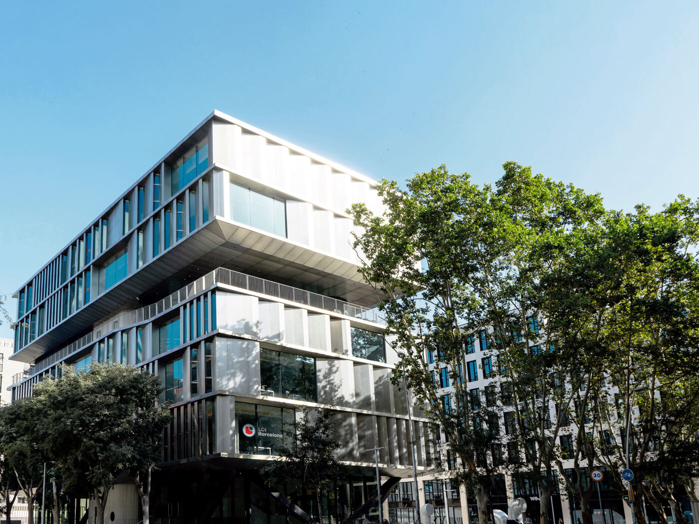A modern, multi-story building with a unique architectural design stands amidst lush green trees under a clear blue sky.