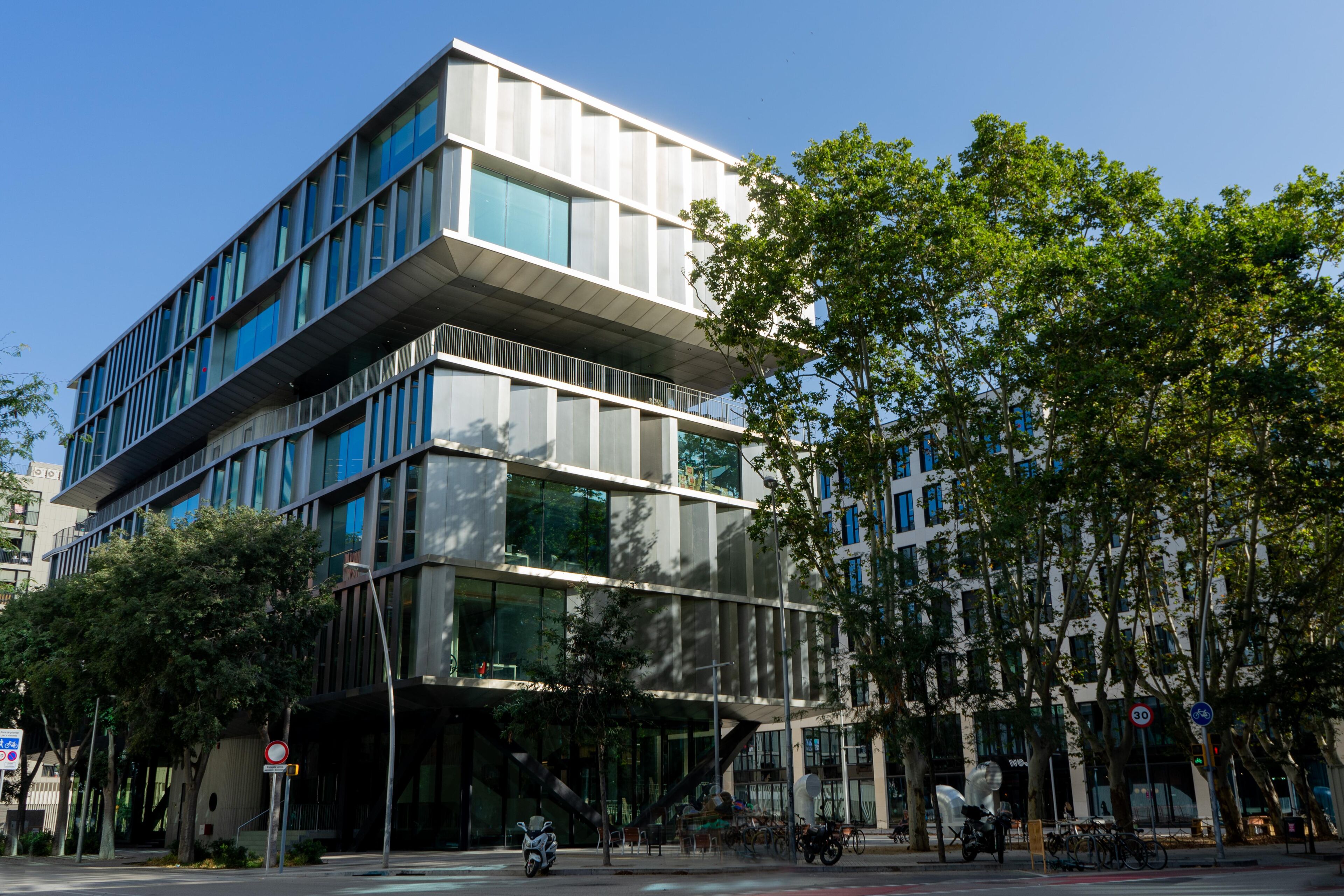 A modern office building with a geometric design and large glass windows. The multi-level structure is surrounded by trees, providing a green and pleasant environment. Bicycles and scooters are parked along the street, adding to the urban scene.