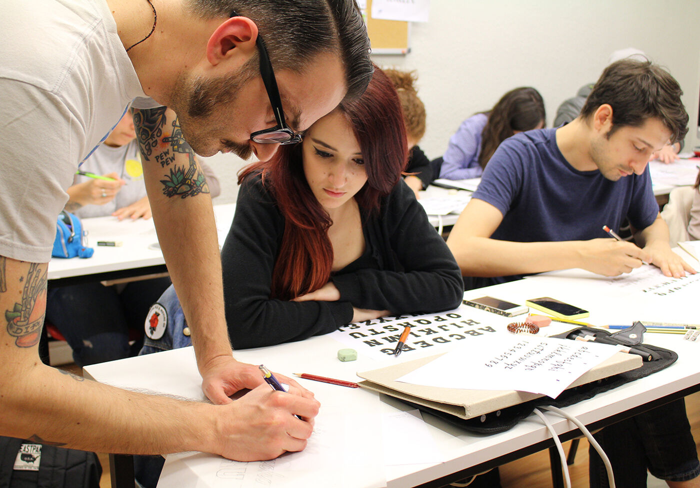 An instructor assists a student during an art class, with peers focused on their work.