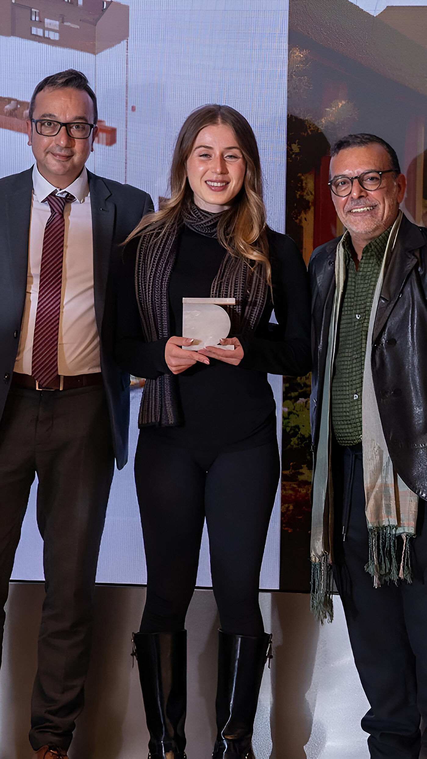 The image shows three people posing for a photo. A woman in the center is holding an award. They are all smiling and appear to be at an event.
