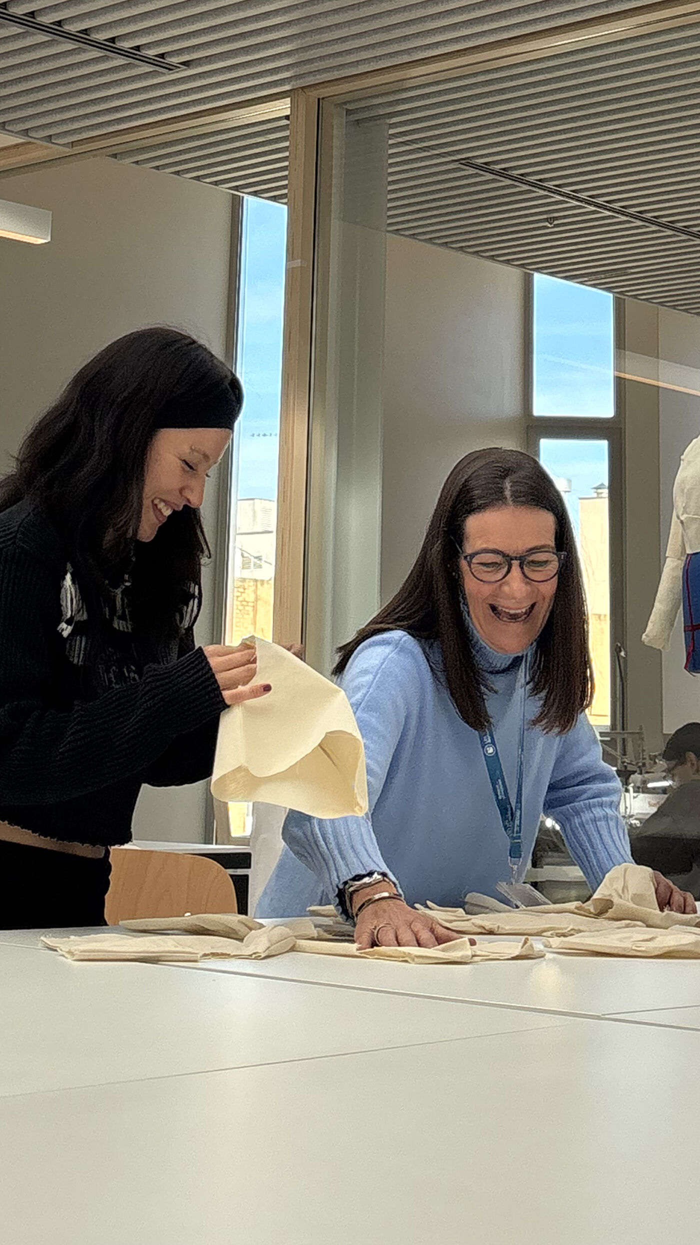 Two women are engaged in a fabric crafting activity in a brightly lit room with large windows. One wears a black sweater and smiles while holding a piece of fabric. The other, in a light blue sweater, laughs warmly. The setting suggests a creative or educational workshop.