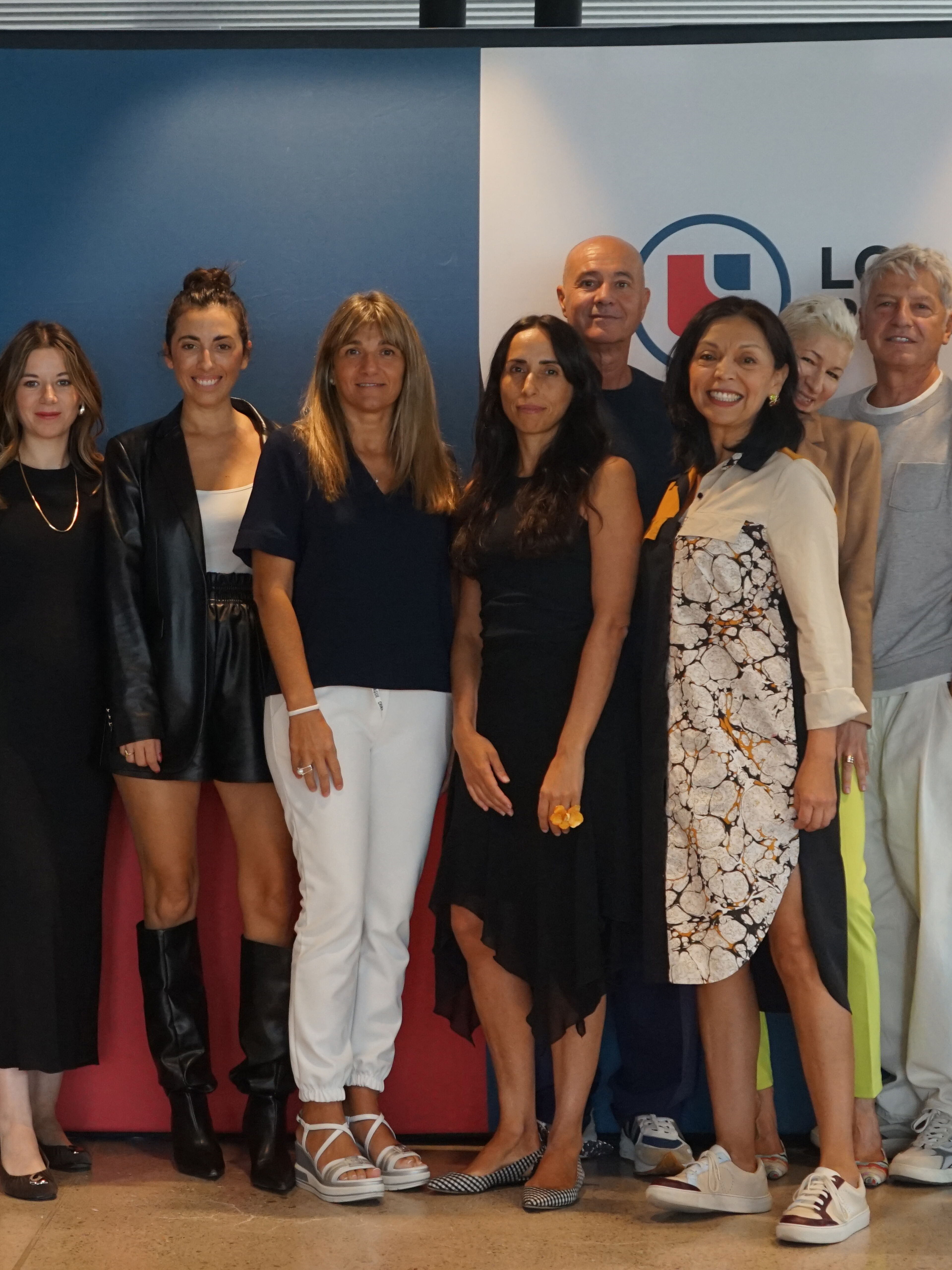 A diverse group of eight men and women stands in front of a branded backdrop, posing for a professional team photo. They wear a mix of business casual and trendy attire, smiling warmly at the camera, suggesting a collaborative and friendly work environment.