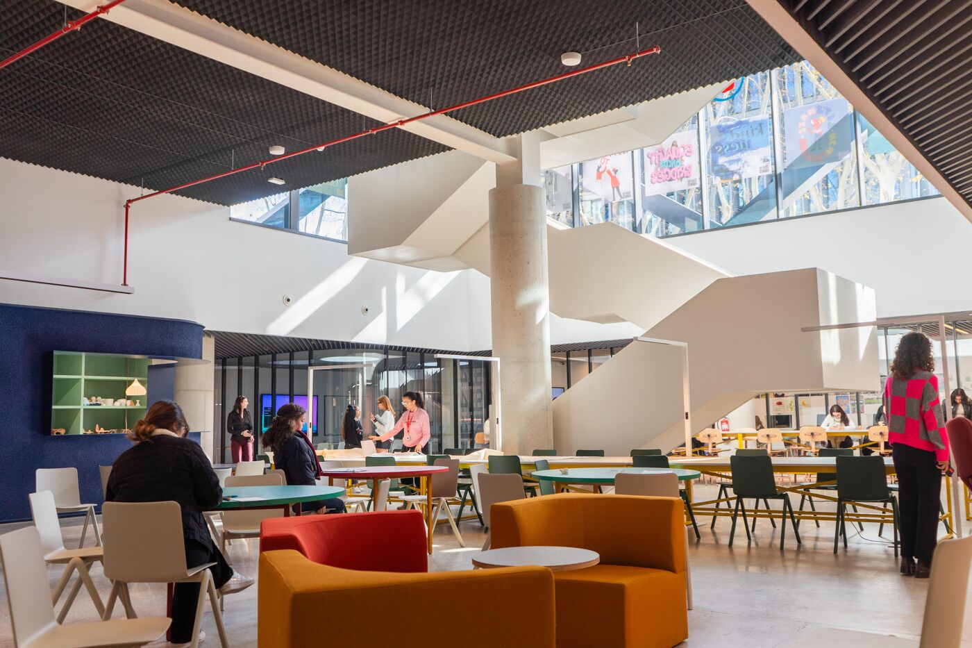 A bright, modern interior space with a large, white spiral staircase, colorful furniture, and people socializing and working at tables. The space appears to be a co-working area or a university common room.