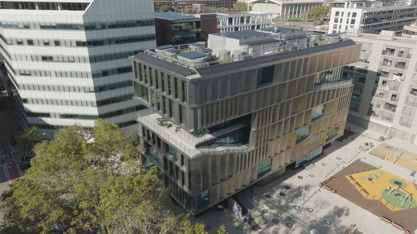 High-angle, long shot of a modern, angular building situated in a city setting. 


The building is multi-storied with a textured, bronze-colored facade. It features several levels of balconies or terraces and a rooftop that seems partially covered by solar panels. The building's design is unconventional, with angled walls and recessed sections. 


Surrounding the main building are other structures, including taller, sleeker buildings with glass facades, and shorter, more traditional-looking buildings. There's also a significant amount of green space visible, including mature trees and a landscaped area that appears to include playground equipment. The ground around the buildings is paved, showing a square or plaza-like design. The overall lighting suggests it's daytime, with bright, natural light illuminating the scene.