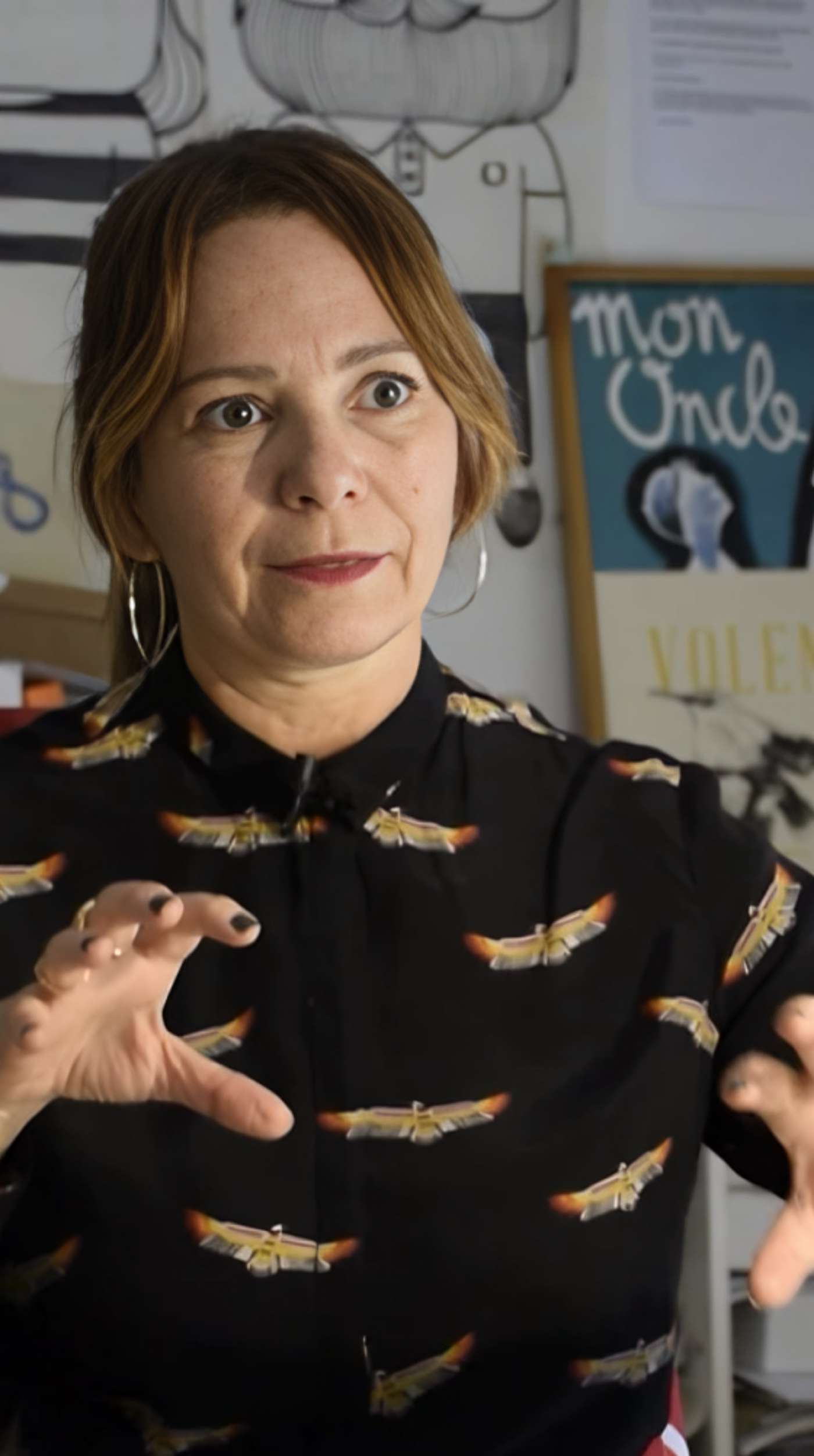 A woman passionately explains a concept in a creative workspace adorned with posters, illustrations, and books. Her expressive gesture and attentive gaze convey enthusiasm and engagement in a discussion or presentation.