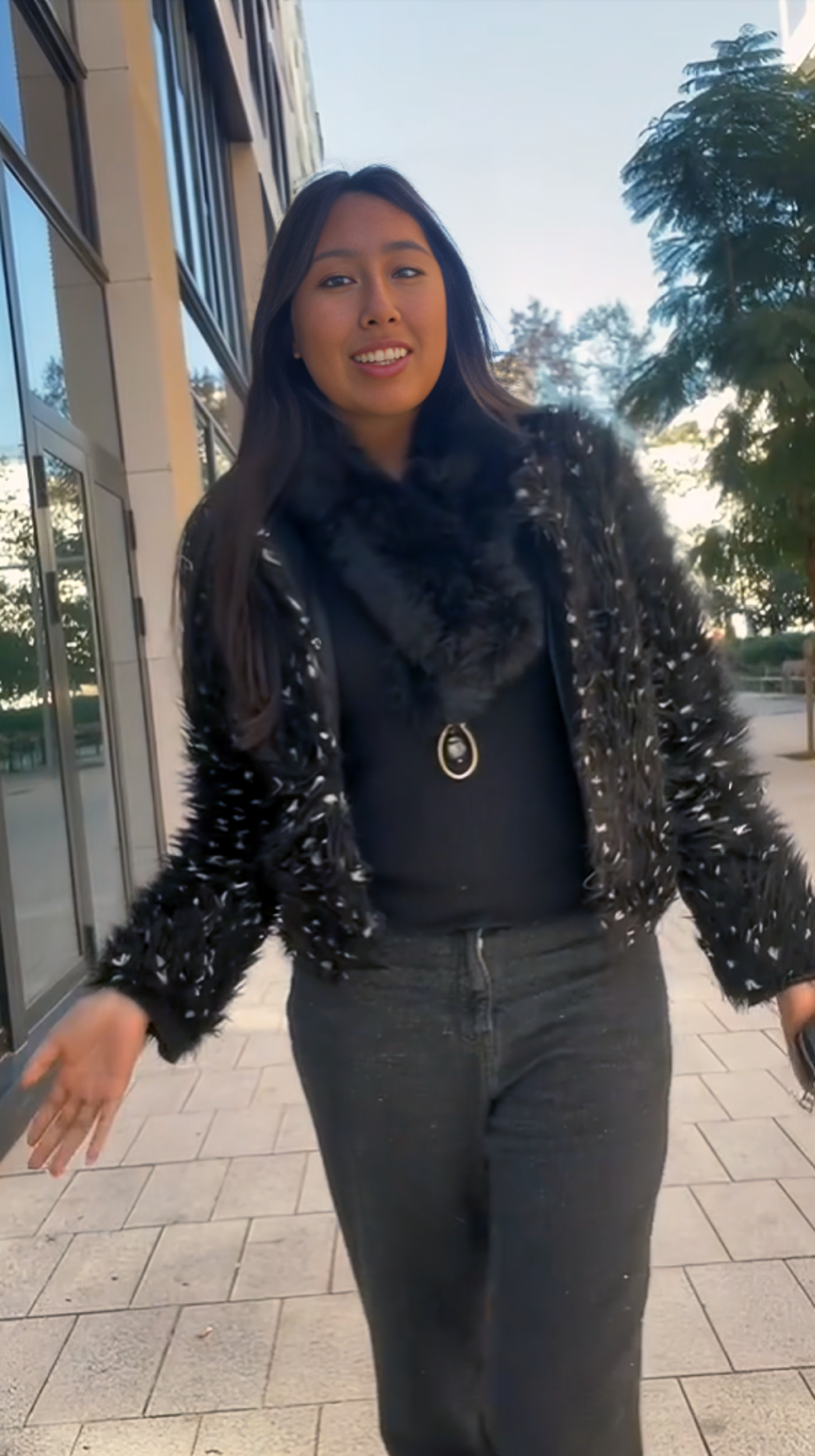 A smiling woman wearing a black fur-trimmed jacket, a statement necklace, and dark jeans walks confidently in an outdoor urban setting. The modern background with trees and buildings complements her chic and fashionable appearance.