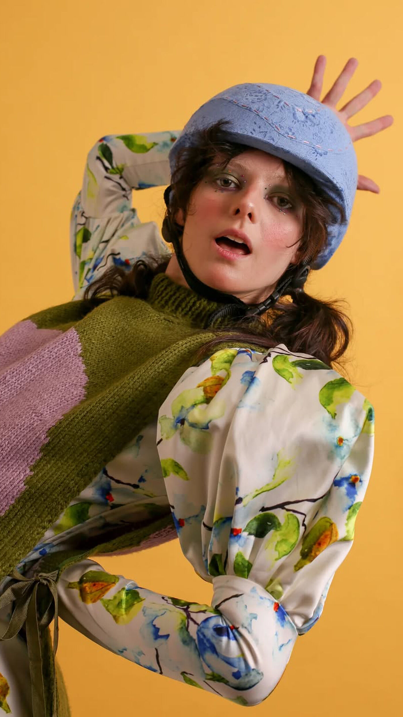 A woman with fair skin and dark hair poses in front of a yellow background. She is wearing a blue helmet, a floral-patterned blouse, and a colorful sweater. Her right hand is raised, fingers spread, near her head.