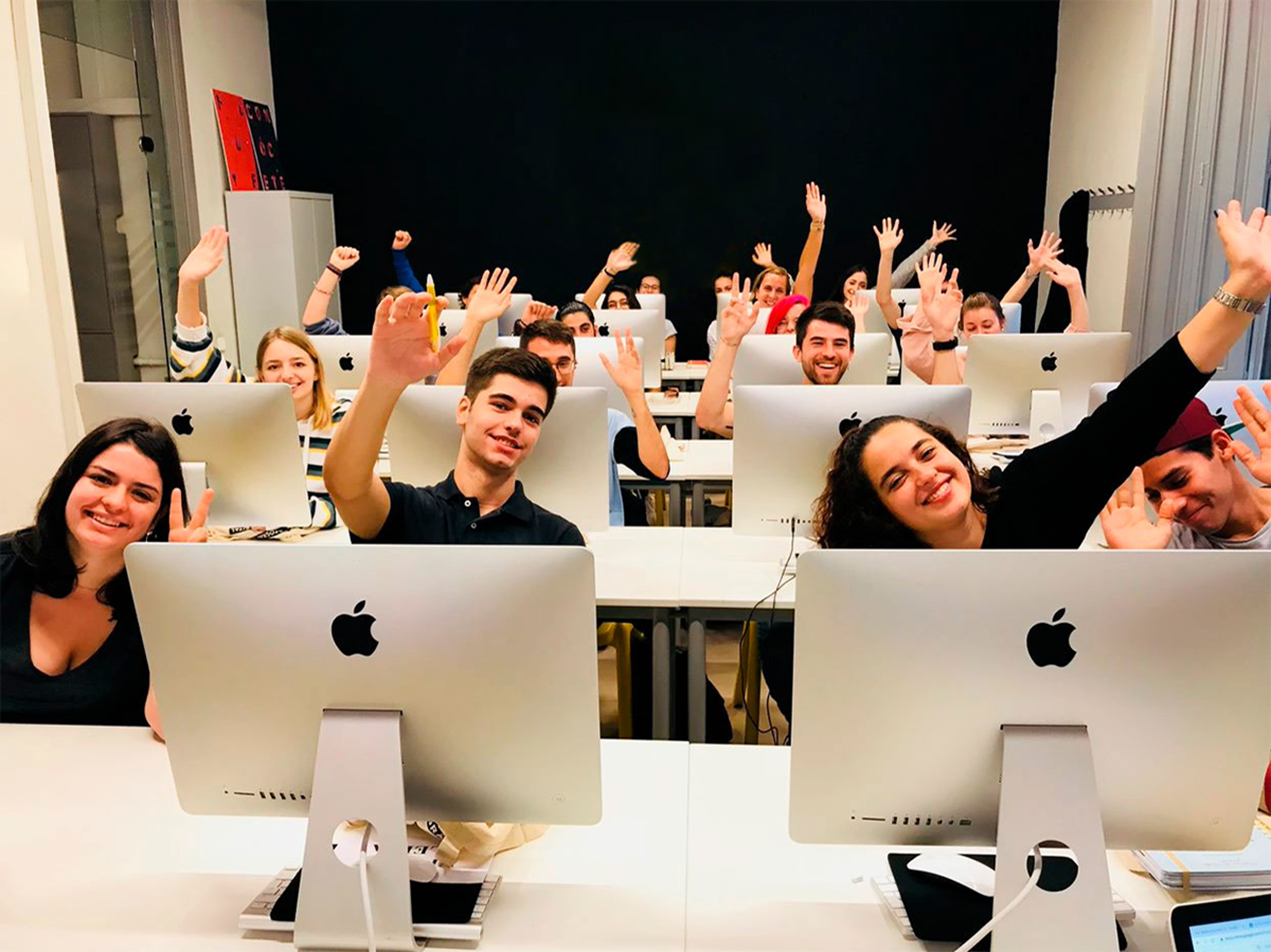 Students waving and smiling behind computer monitors in a modern computer lab, exhibiting enthusiasm and positivity.