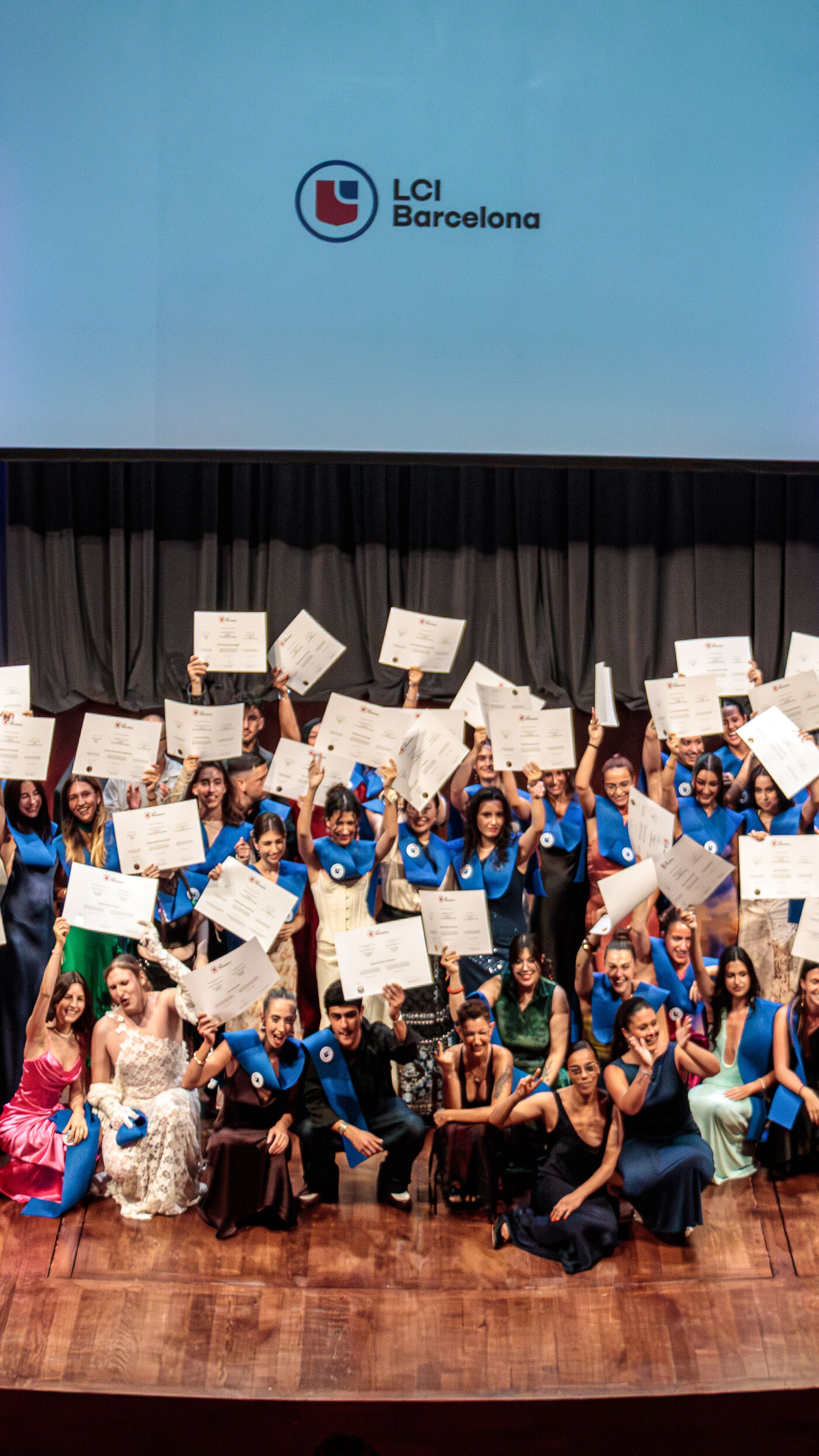 A group of graduates from LCI Barcelona are on stage, holding up their diplomas and celebrating their achievement. They are dressed in graduation attire, and the LCI Barcelona logo is visible in the background.