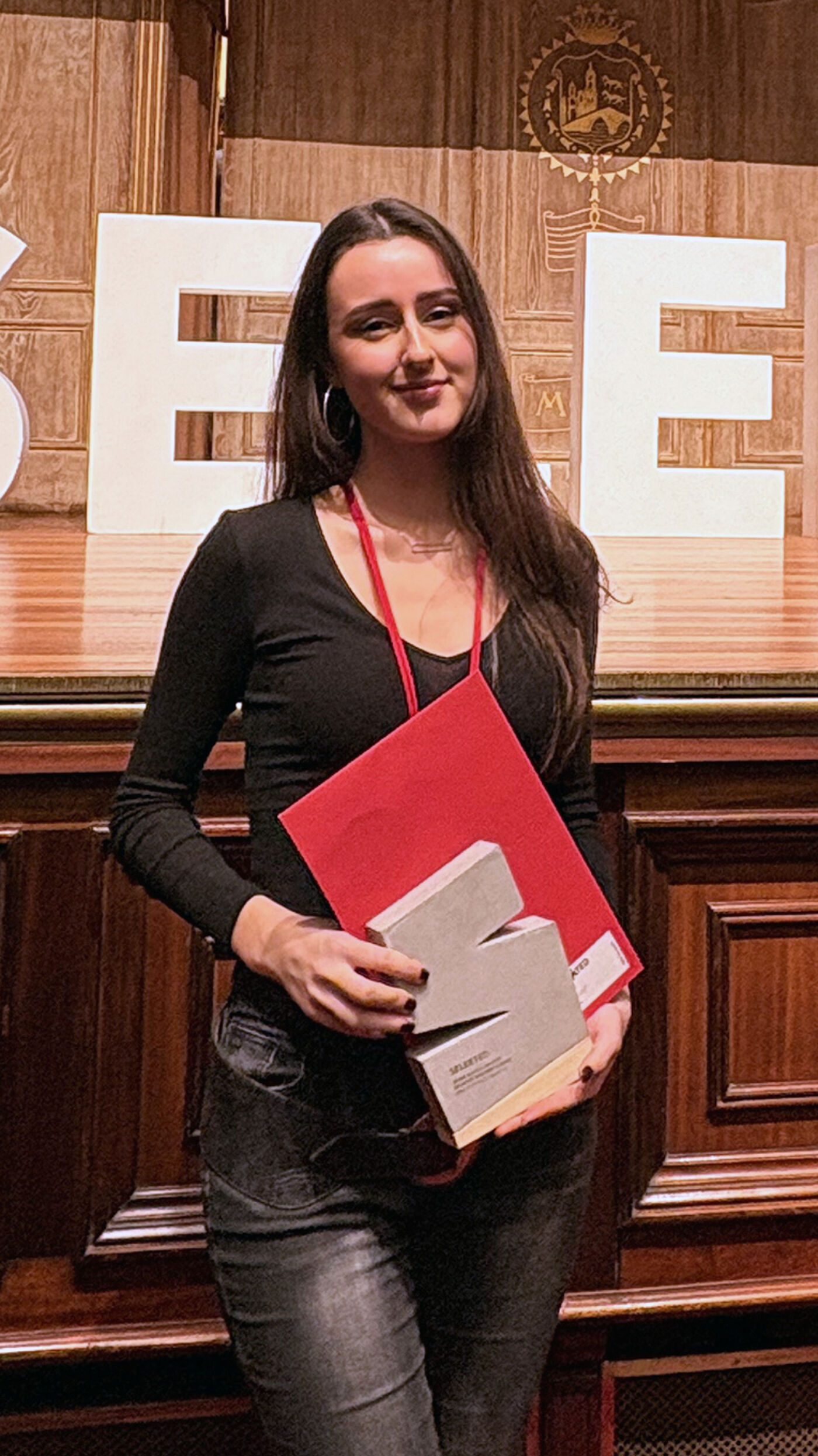 A smiling woman with long, dark hair holds an award and a red folder. She is standing in front of a large sign with the letter 'E'.