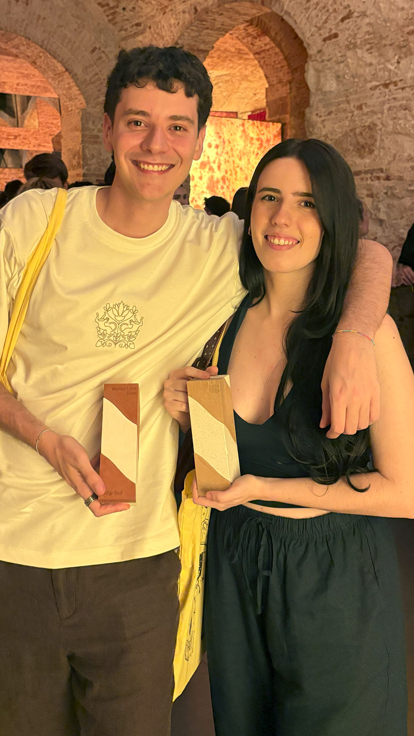 A smiling man and woman are posing indoors, each holding a small, rectangular object with a diagonal design. The man is wearing a light-colored t-shirt and dark pants, while the woman is in a dark top and pants. They appear to be at an event or gathering.