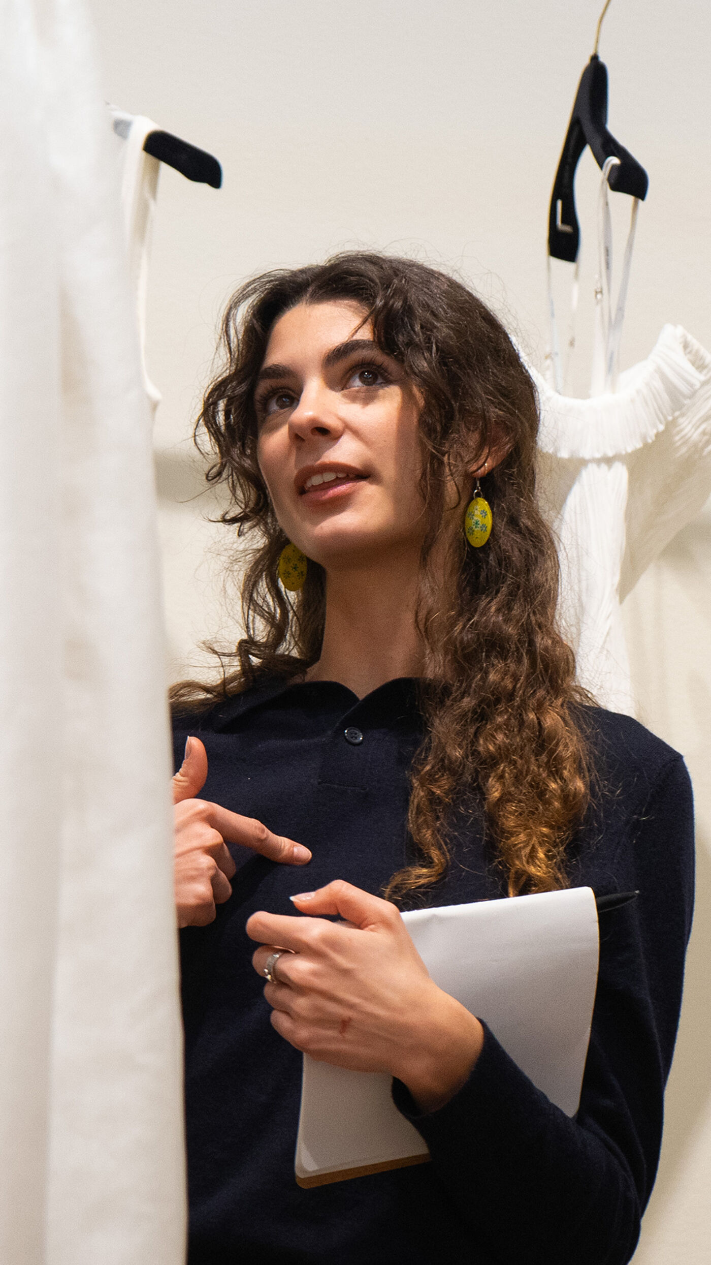 A fashion designer, holding a notepad, gestures while discussing design elements in a workspace. Surrounded by white garments on hangers, her expressive demeanor highlights a focus on creativity and collaboration in the fashion industry.