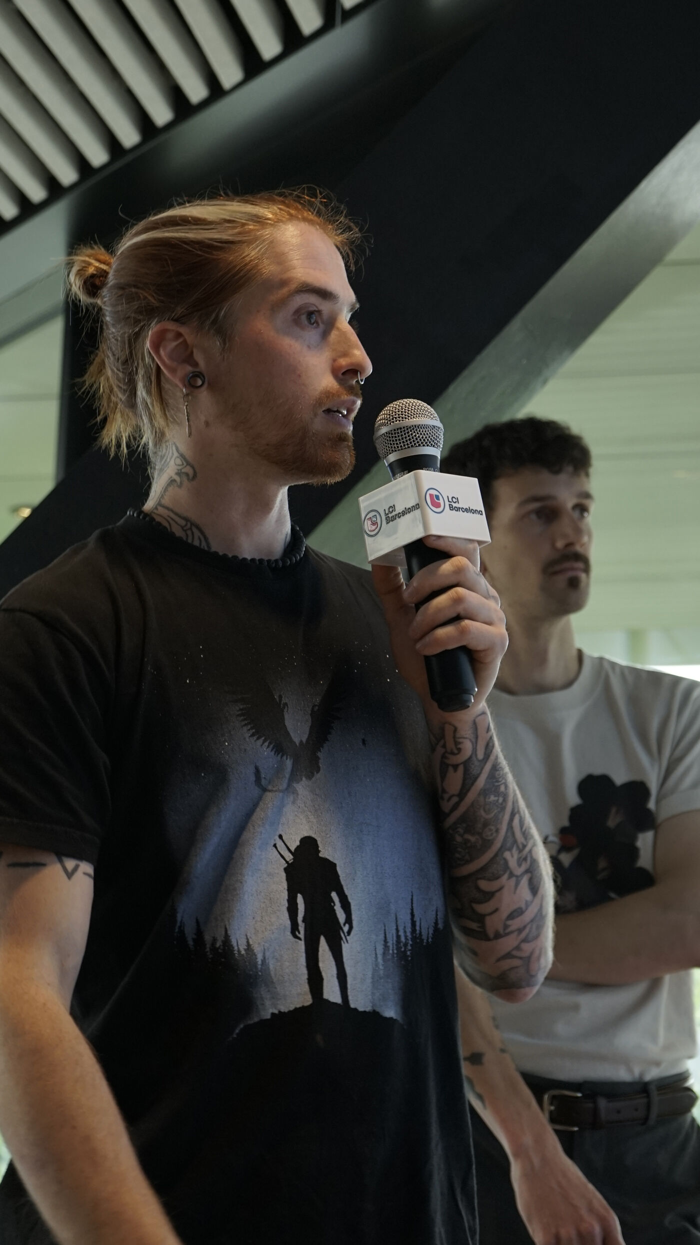 A man with a long ponytail and tattoos speaks into a microphone while another man stands beside him.