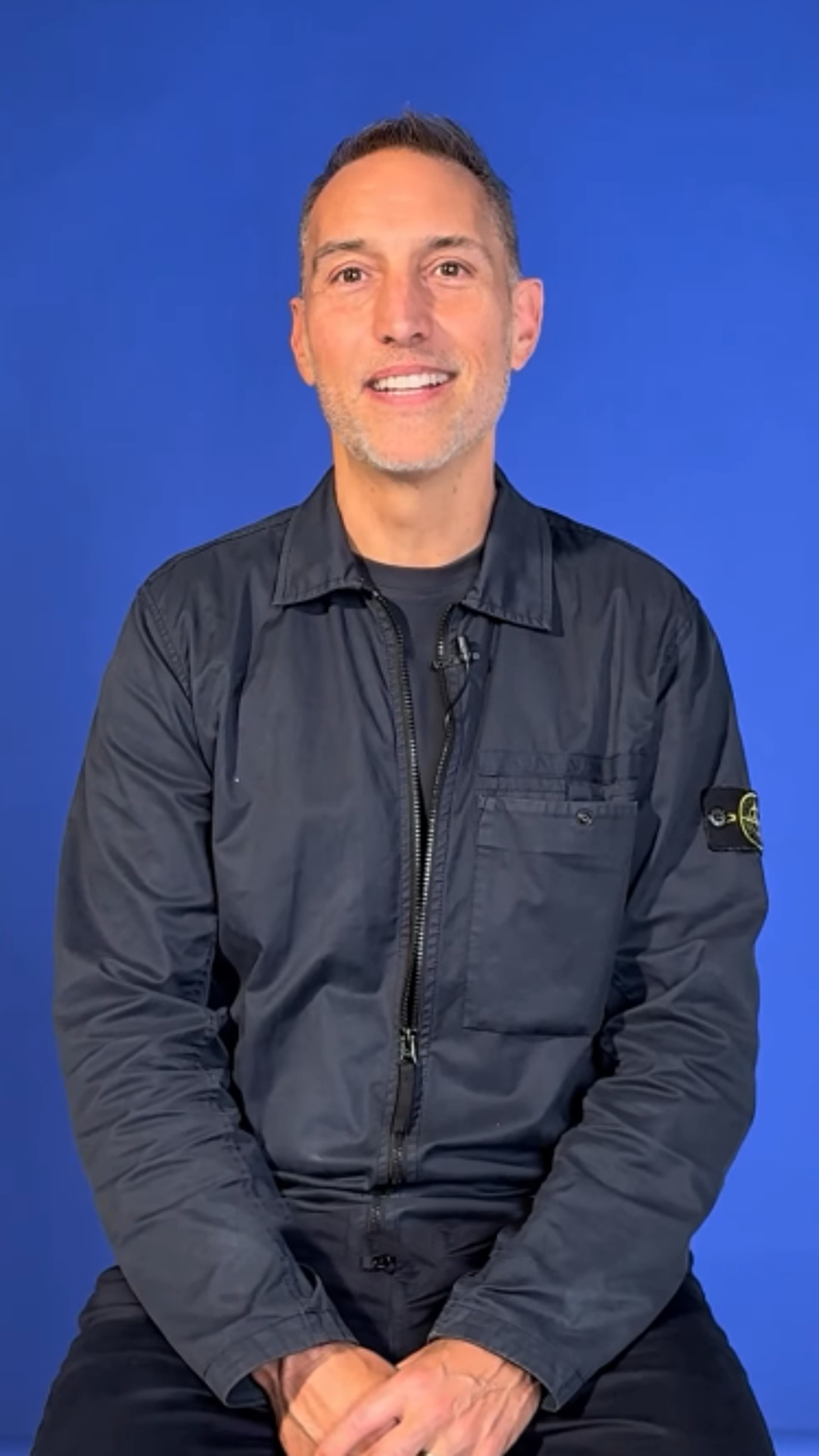 The image shows a man with short, graying hair and a light complexion, wearing a dark blue jacket and pants. He is sitting with his hands clasped in front of him, smiling slightly against a solid blue background.