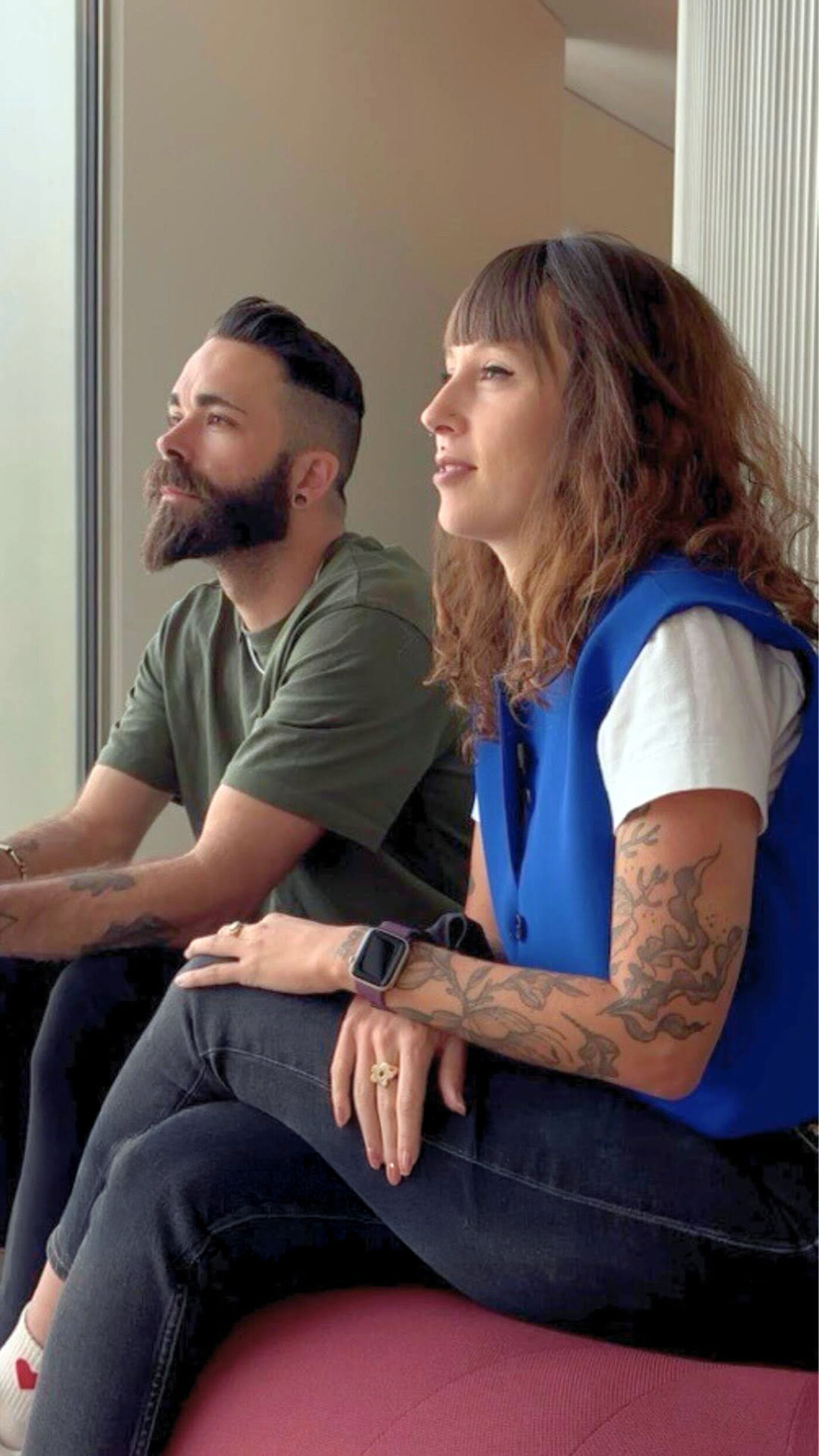 Two people, a man with a beard and a woman with tattoos, sit side-by-side looking out a window.