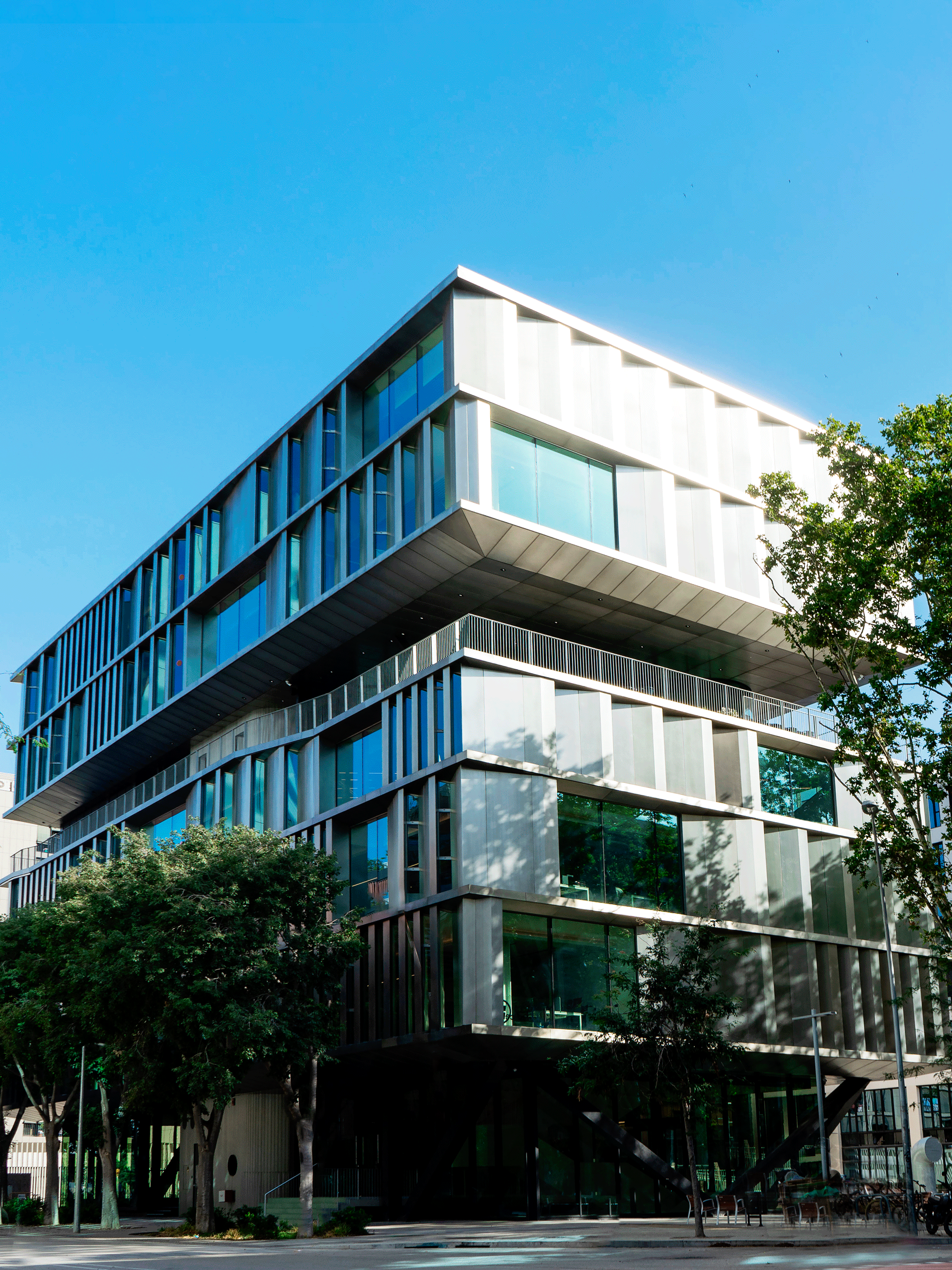 A contemporary multi-story office building with a unique, staggered design and large glass windows reflecting the sky. The structure features alternating layers of glass and metal panels, creating a sleek, dynamic appearance. Trees line the surrounding sidewalk, adding a touch of greenery.