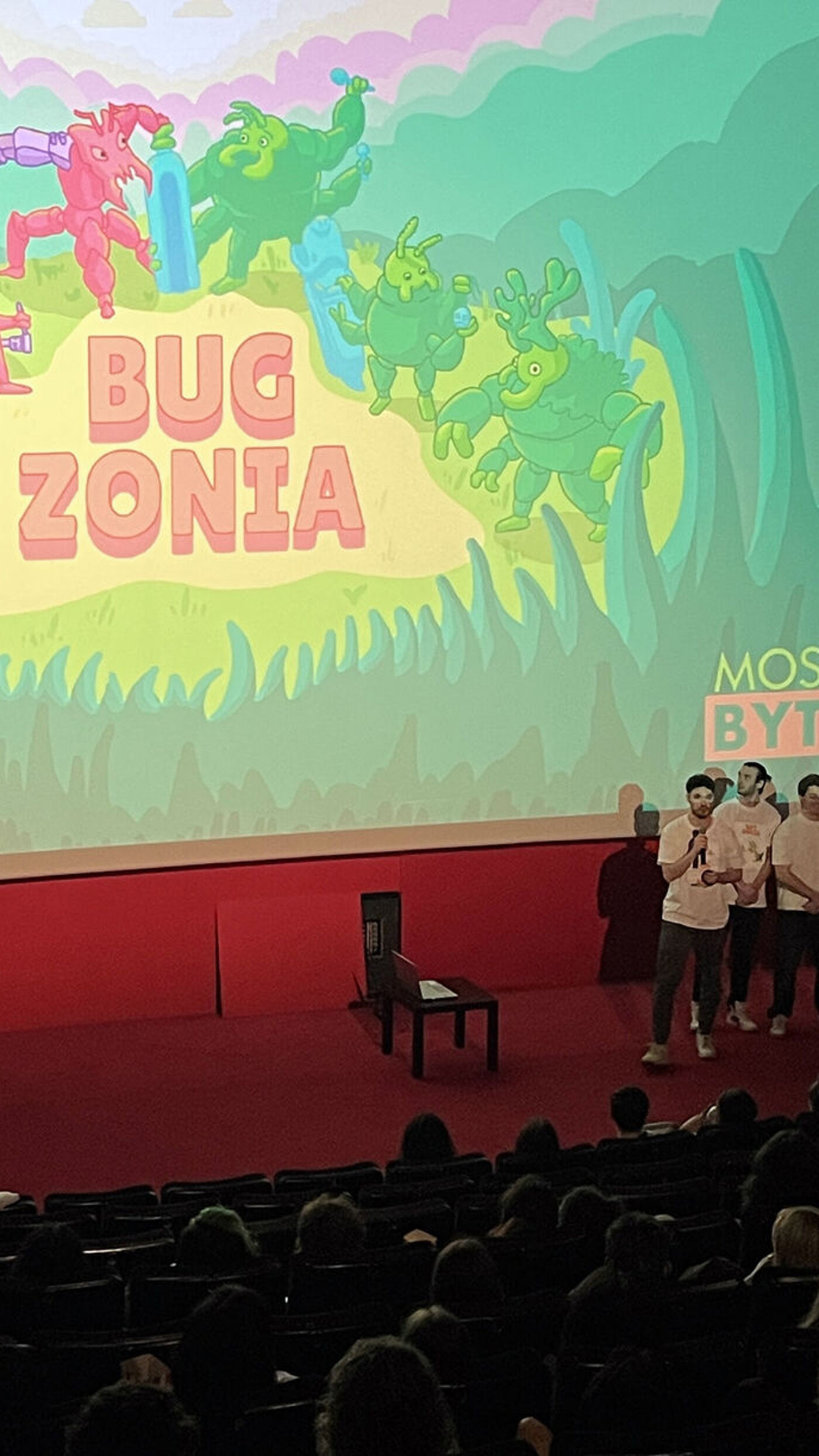 A presentation is being given in a theater with a screen displaying the title "Bug Zonia" and cartoonish characters. A group of people are on stage, addressing an audience seated in the theater. The scene suggests an event related to animation or a children's program.