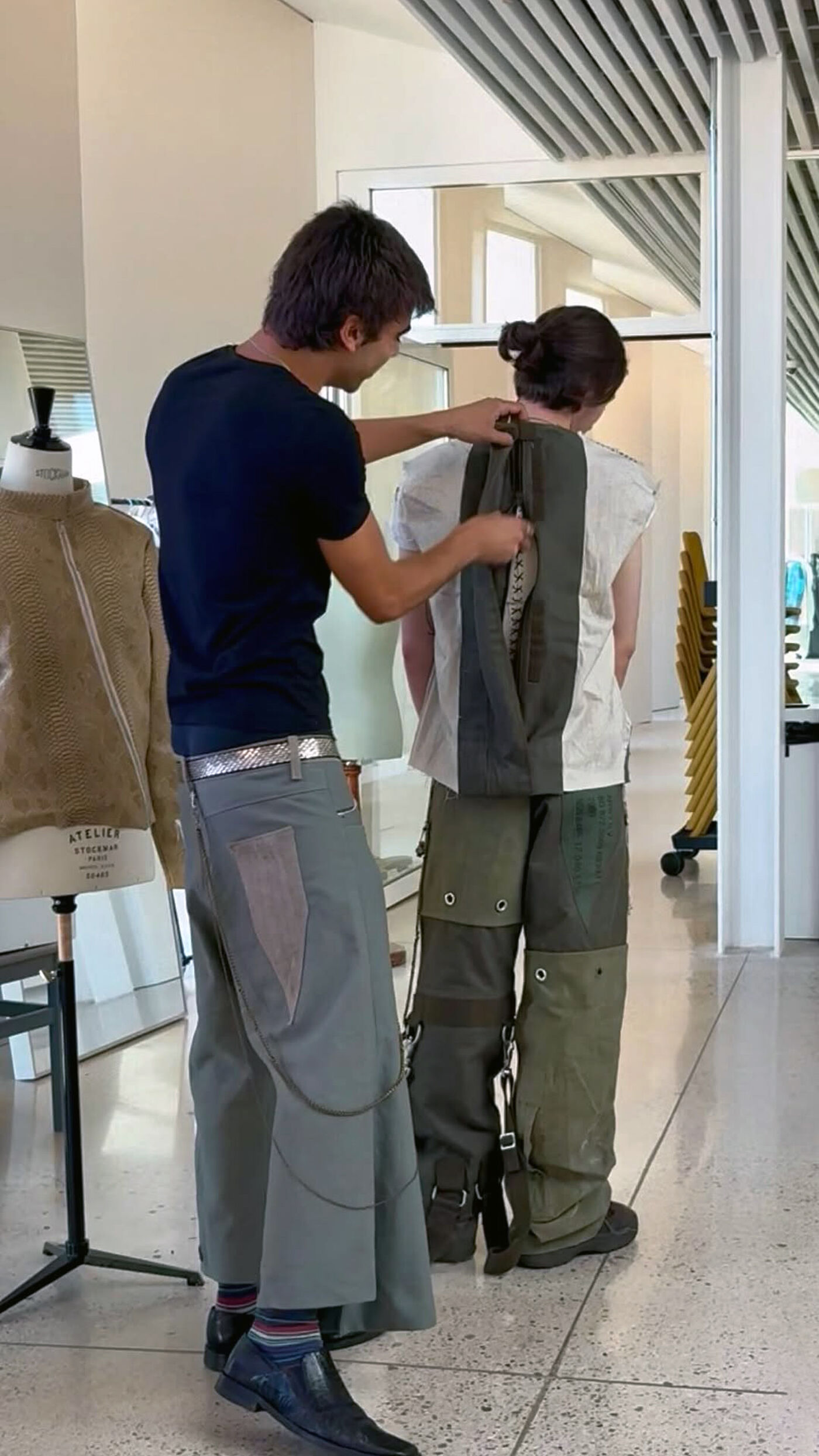 A young man helps another person put on a complex garment in a fashion design studio.