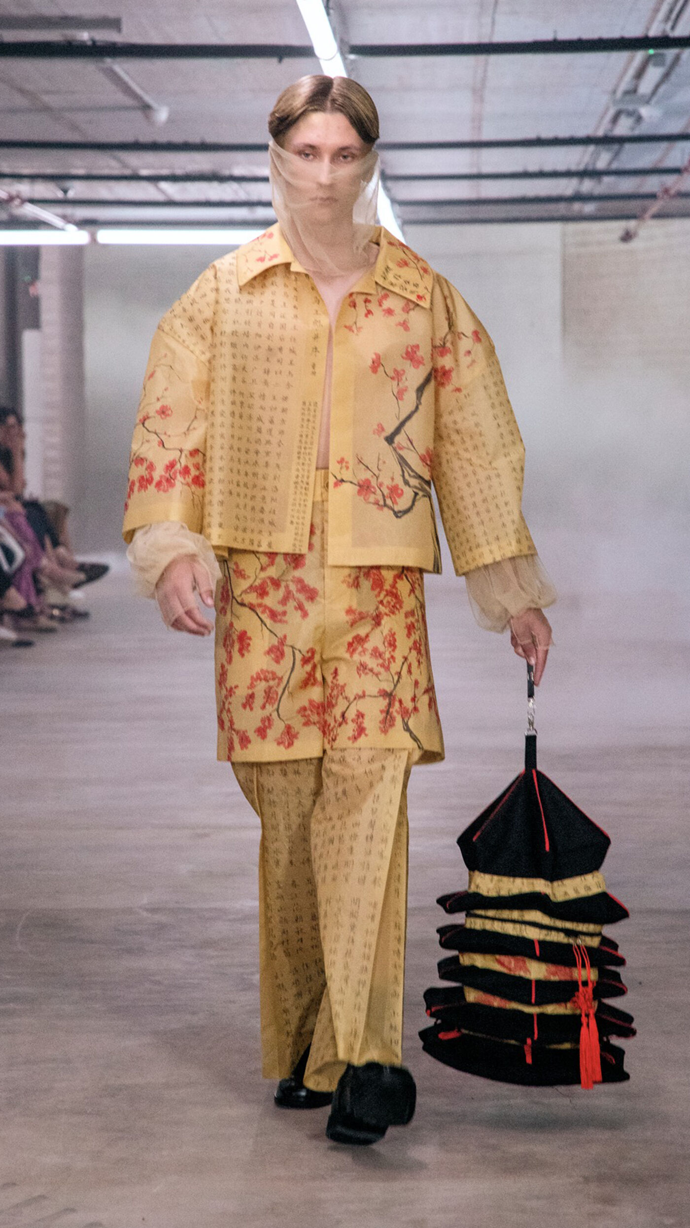 A person walks a runway in a yellow floral patterned suit, holding a tiered black and gold pagoda-shaped object. The outfit includes a jacket, pants, and a sheer head covering, showcasing a unique and artistic design.