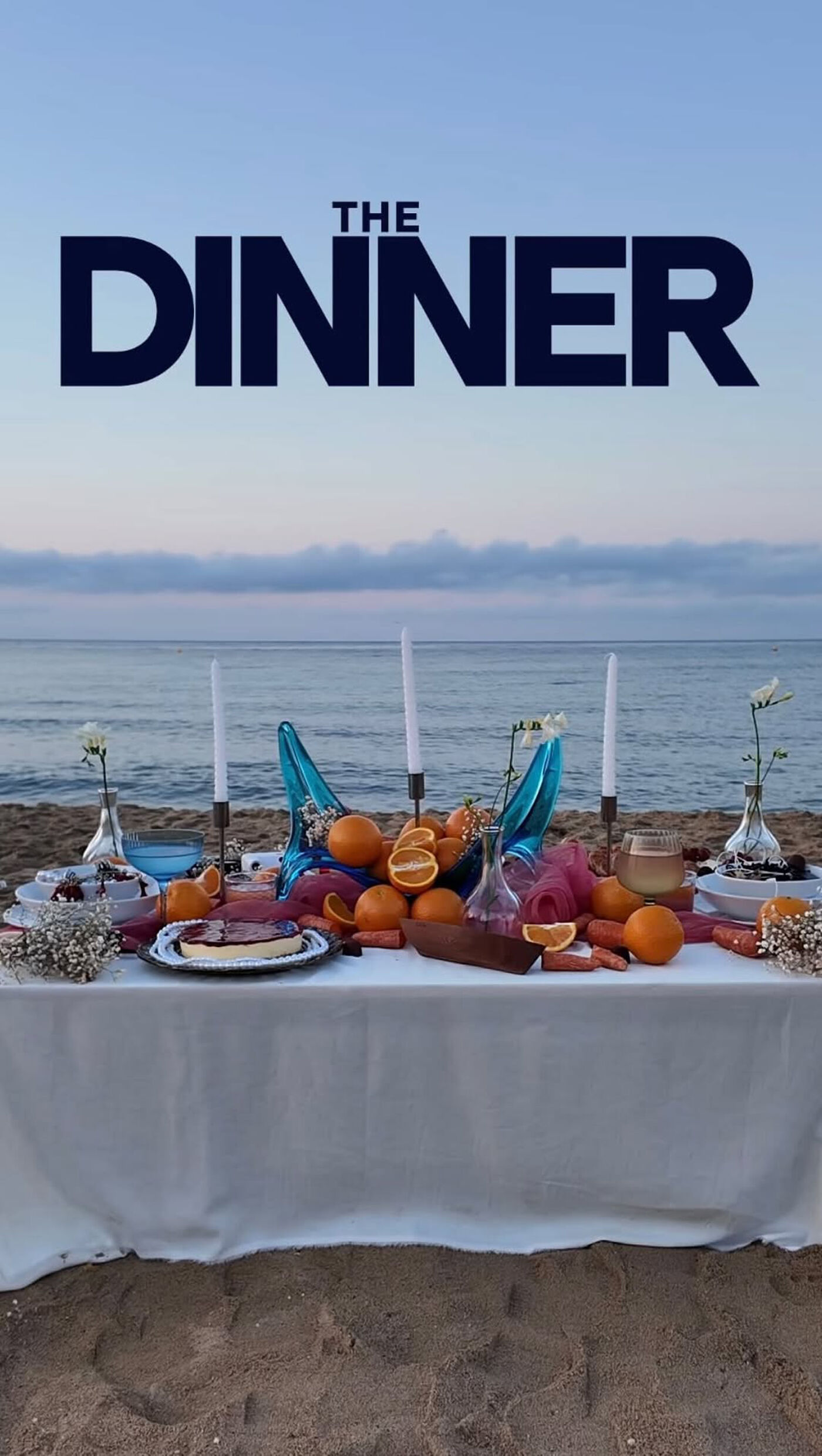 The image shows a beautifully set dinner table on a beach at dusk. Oranges and candles adorn the table, creating a warm and inviting atmosphere against the backdrop of the ocean.