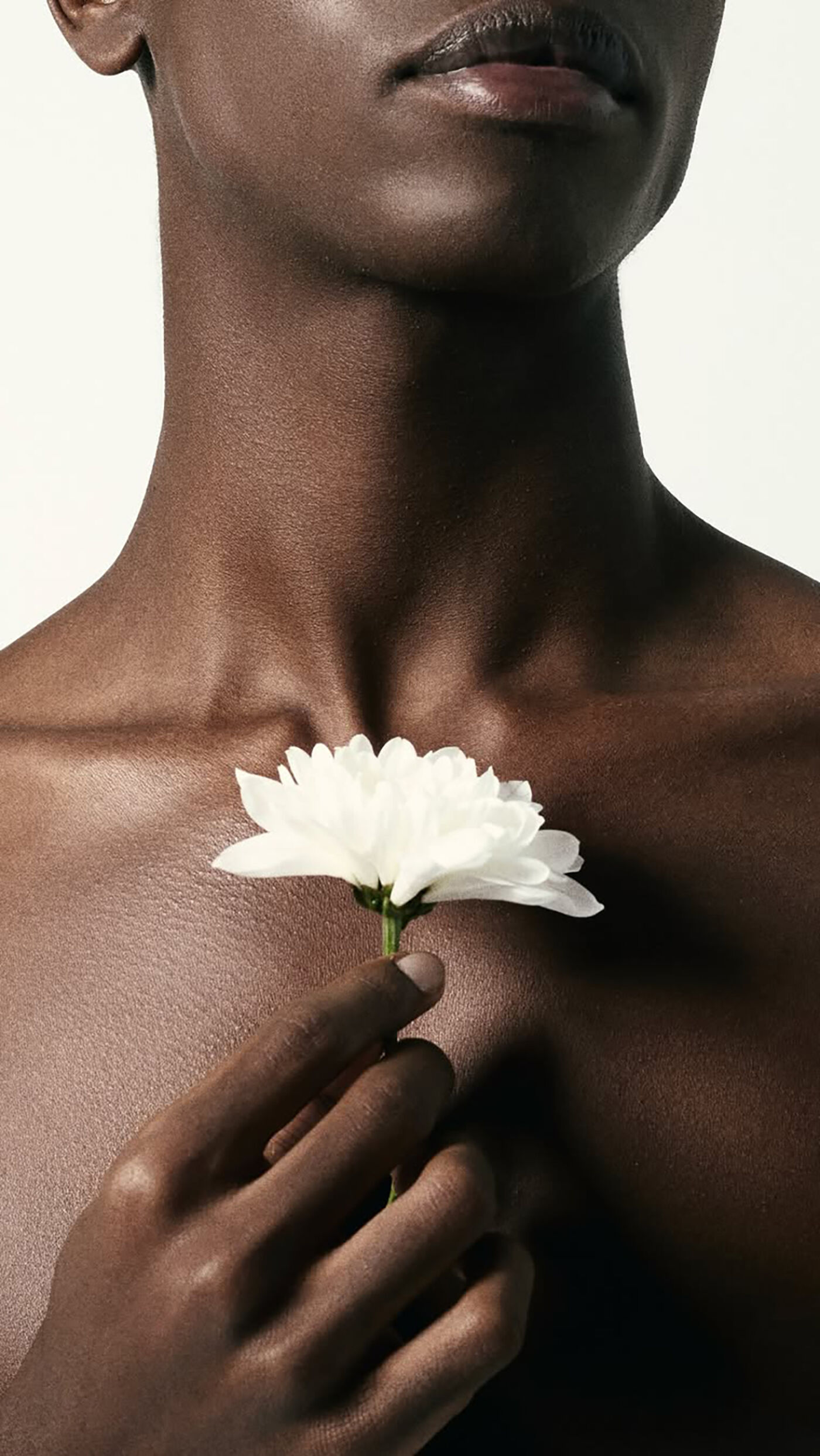 A close-up shot features a person with dark skin holding a white flower against their chest. The focus is on the contrast between the skin tone and the flower's color, highlighting the flower's delicate petals.