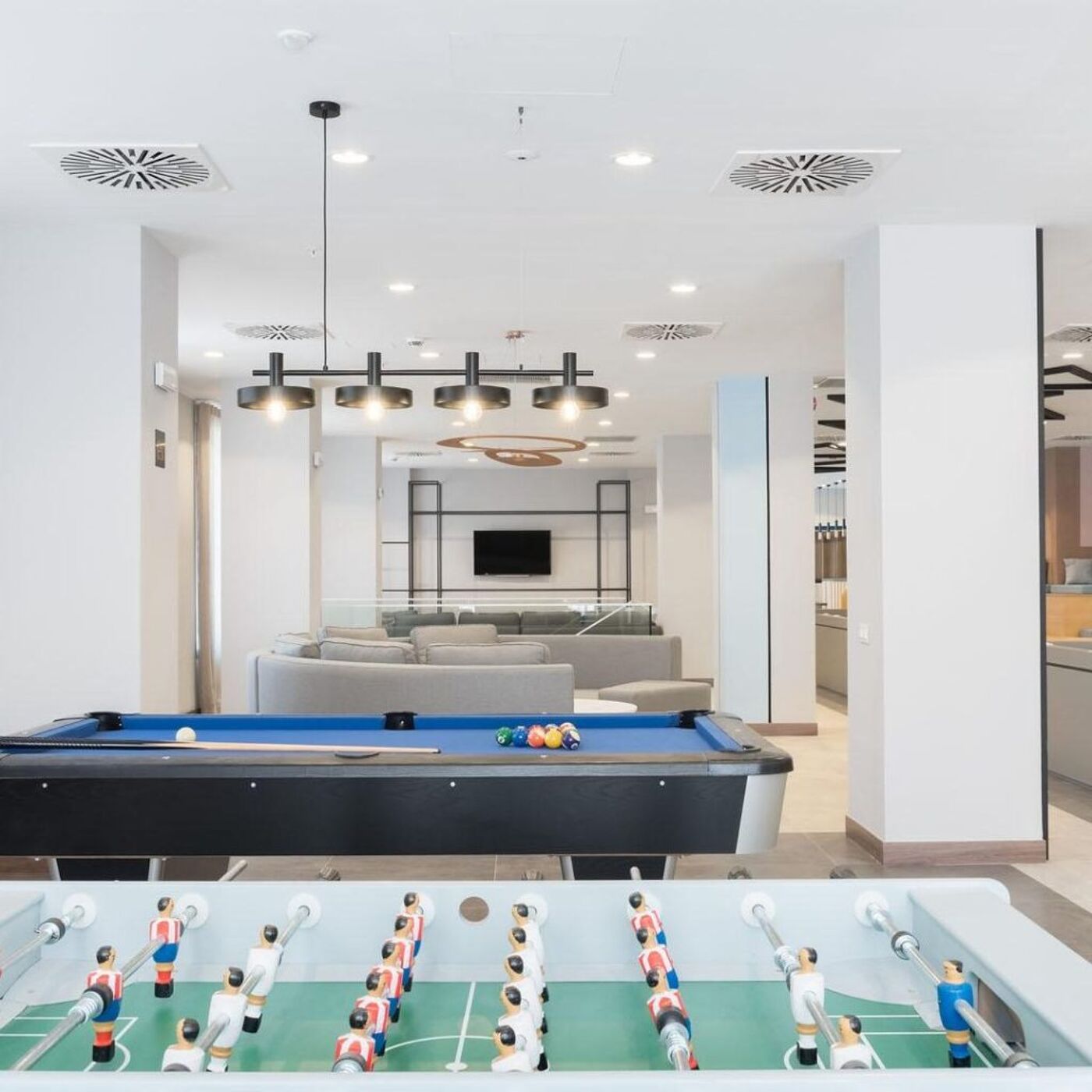 A spacious and modern lounge featuring a pool table and a foosball table in the foreground. The room is well-lit with sleek pendant lights, comfortable gray sofas, and a wall-mounted TV in the background, creating a relaxing and recreational atmosphere.