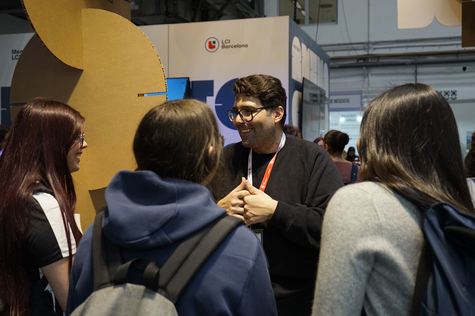 A group of four people are gathered in what appears to be a convention or trade show setting. A man with glasses is speaking to the group, while the others are listening attentively. There's a booth or display in the background.