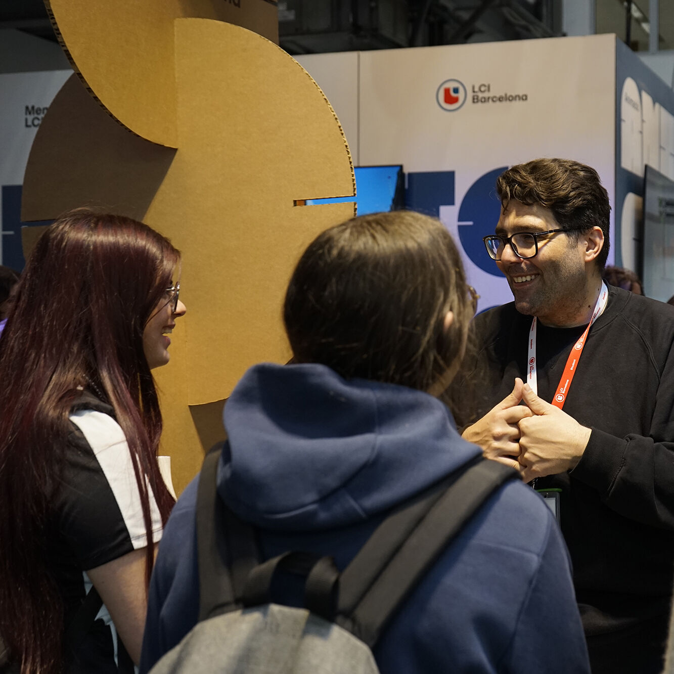 A group of four people are gathered in what appears to be a convention or trade show setting. A man with glasses is speaking to the group, while the others are listening attentively. There's a booth or display in the background.