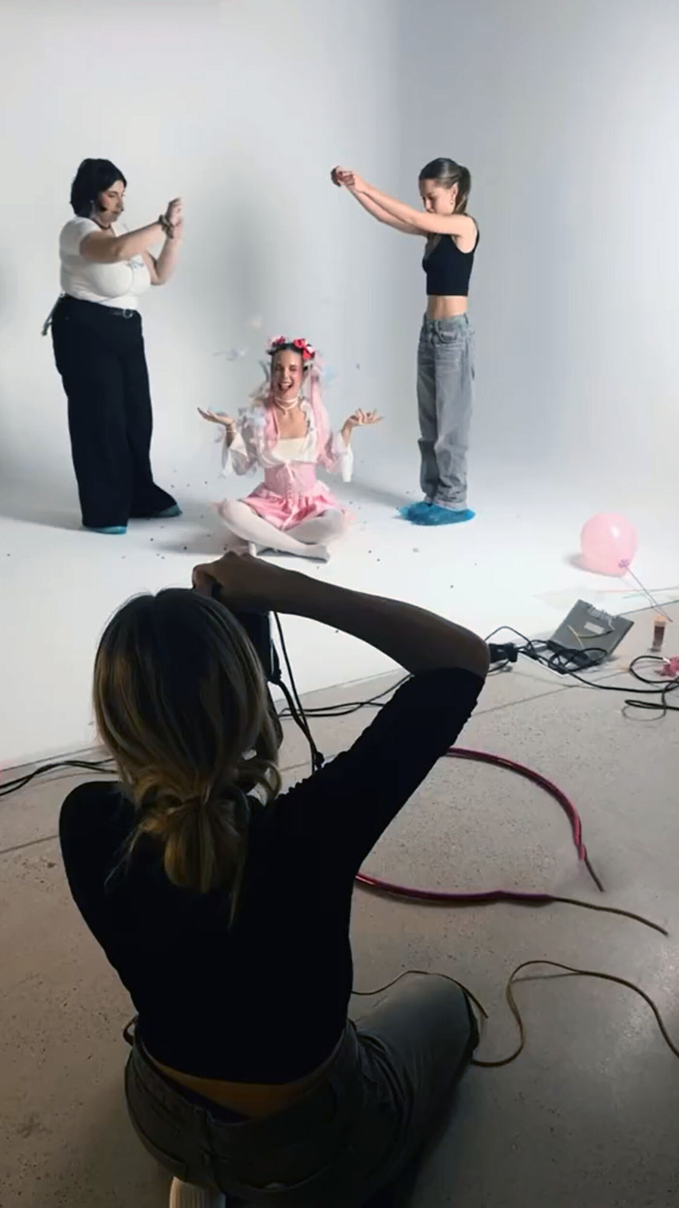 A photographer captures three women posing in a studio, one in a pink dress, one in jeans, and one in black pants, with a pink balloon and camera equipment visible.