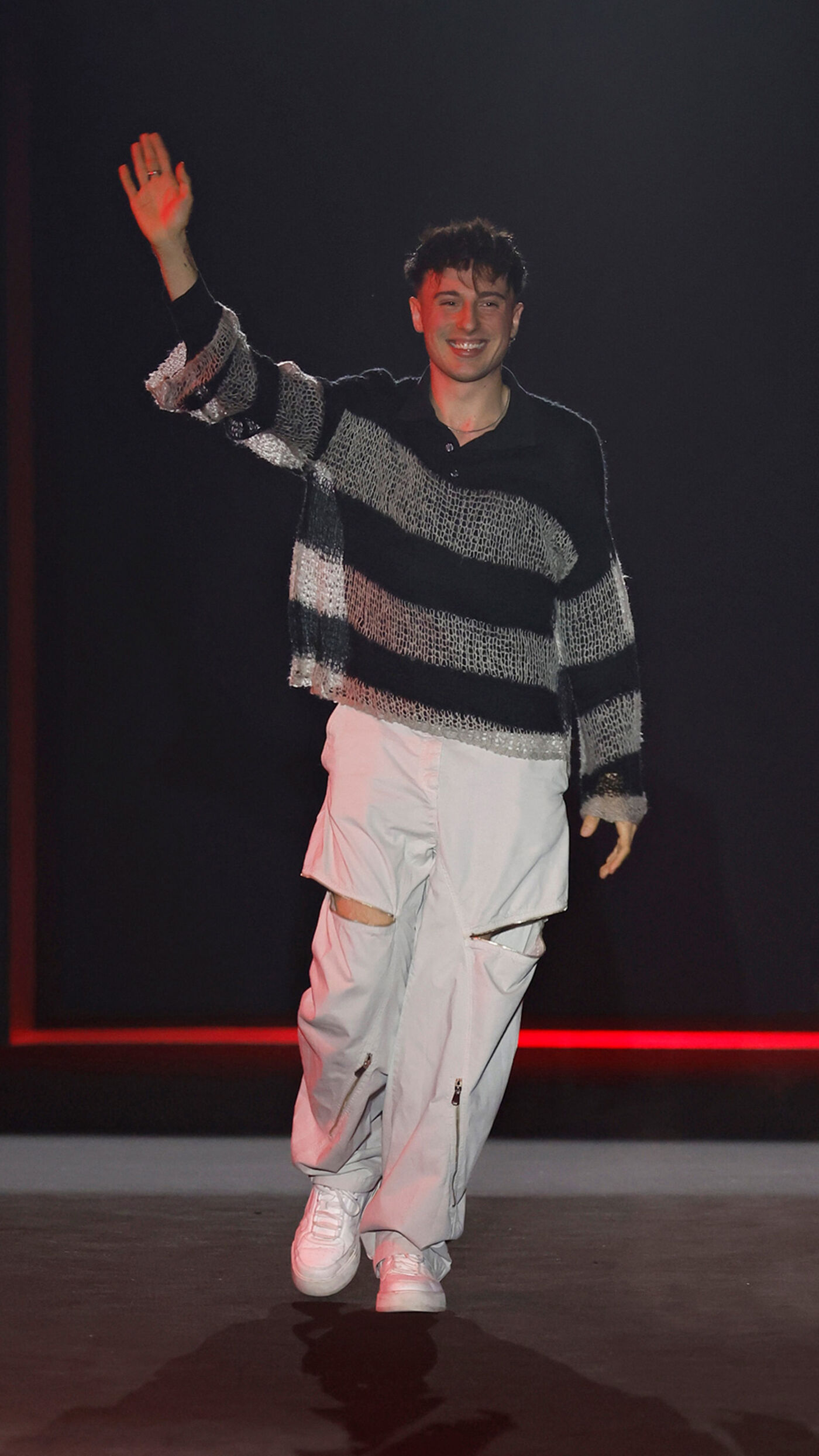 A young man with short brown hair, wearing a black and white striped sweater and light grey pants with rips at the knees, waves to the audience while wearing white sneakers.