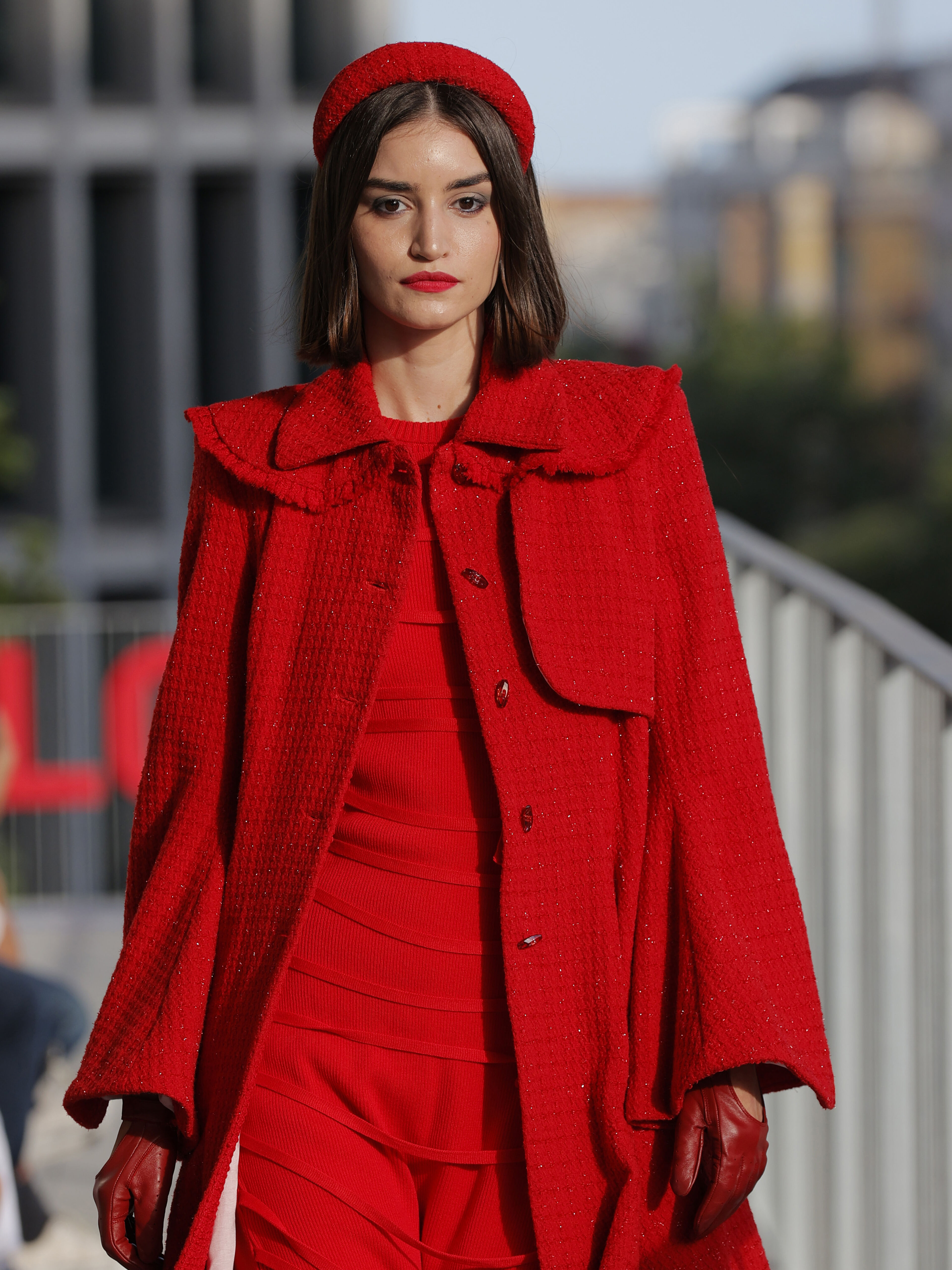  A fashion model walks the runway in a striking all-red outfit, featuring a textured coat, matching dress, gloves, and headband. The bold color choice and elegant style exude confidence and sophistication, capturing attention with a cohesive, monochromatic look.