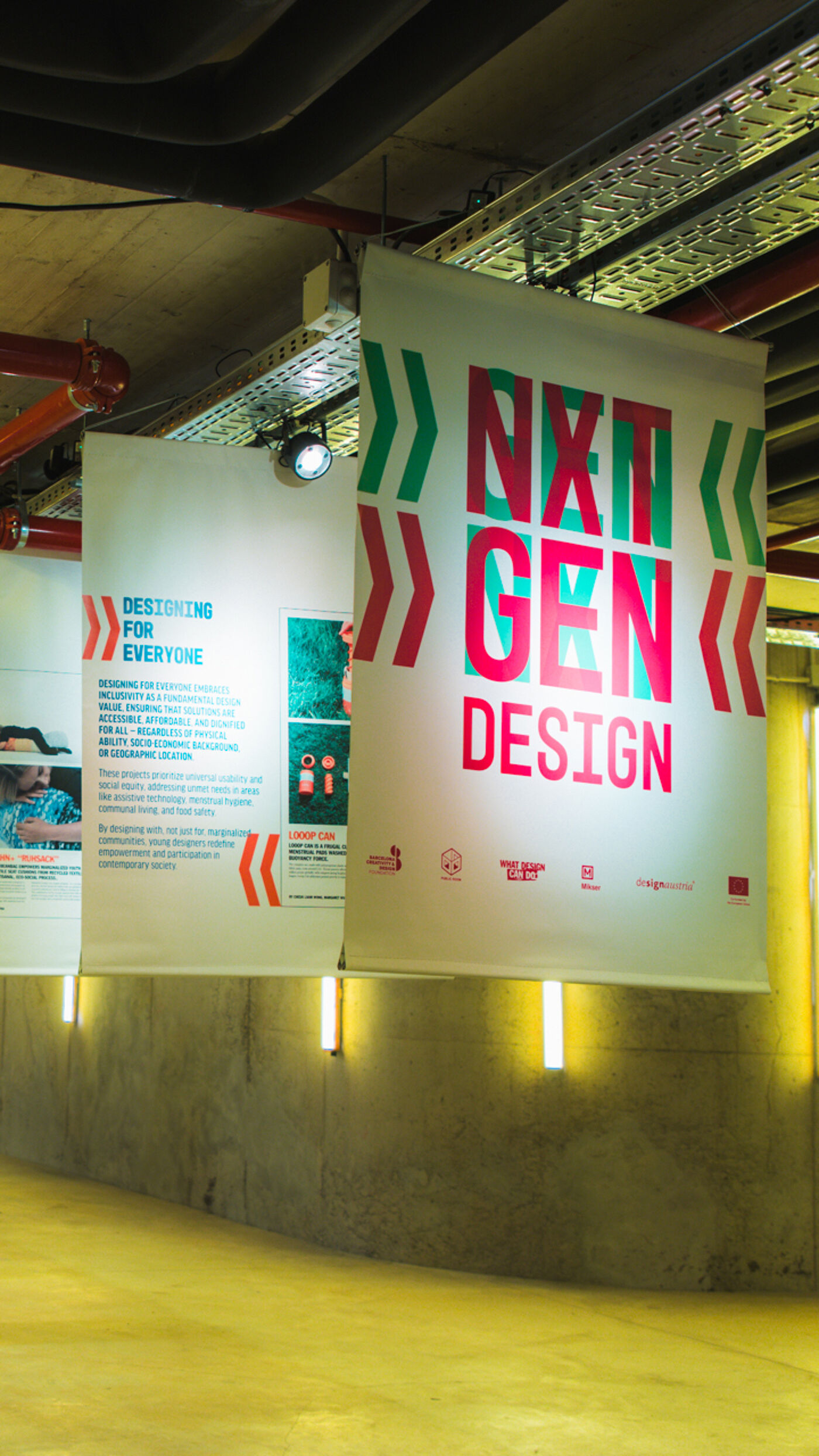 The image shows a banner with the text "NXT GEN DESIGN" in bold, colorful letters. The banner is hanging indoors, likely at a design event or exhibition. There are other banners visible in the background, suggesting a focus on design and innovation.