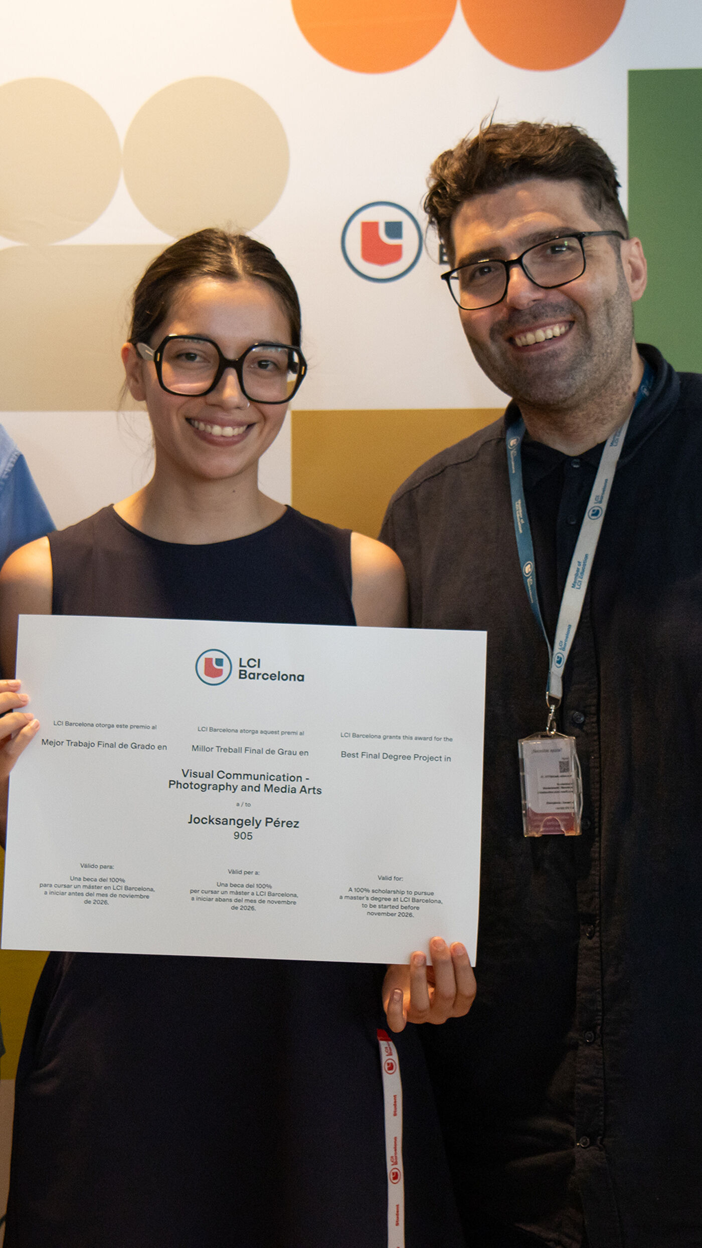 A young woman with glasses holds a certificate, standing next to a man with glasses and a lanyard. They are both smiling in front of a colorful backdrop with circles and a logo.