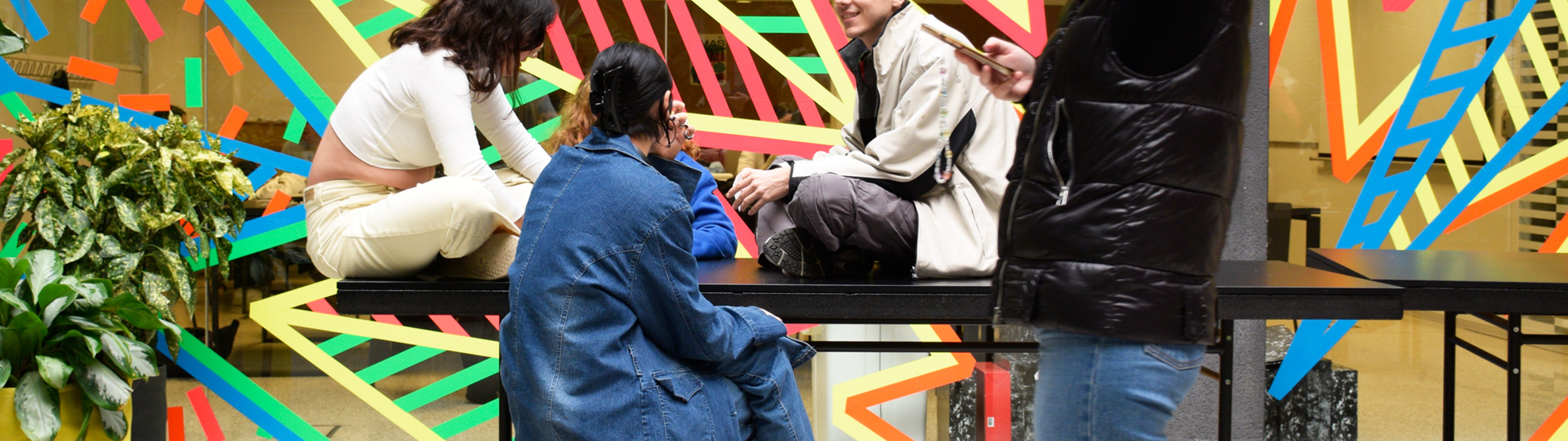 Individuos jóvenes conversando en una zona comunal vibrante y artística con diseños geométricos en la pared.