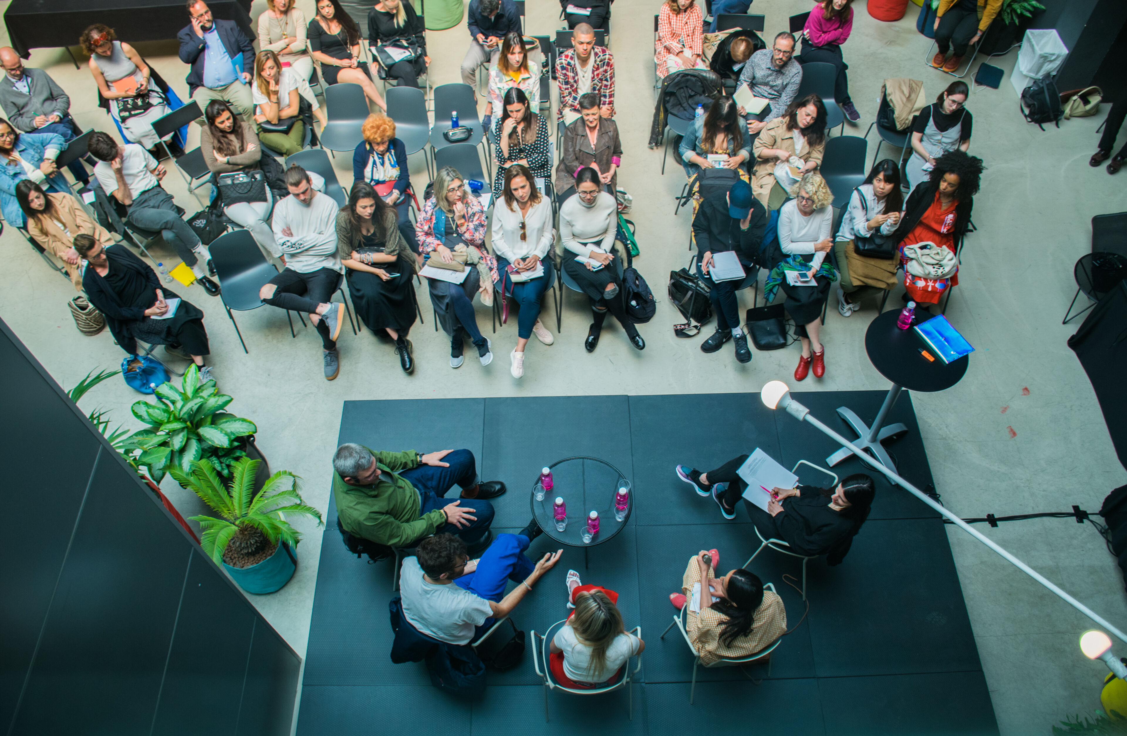 A bird's-eye view captures an engaging workshop in progress, with attendees seated in a semi-circle around the speakers, fostering a collaborative atmosphere.