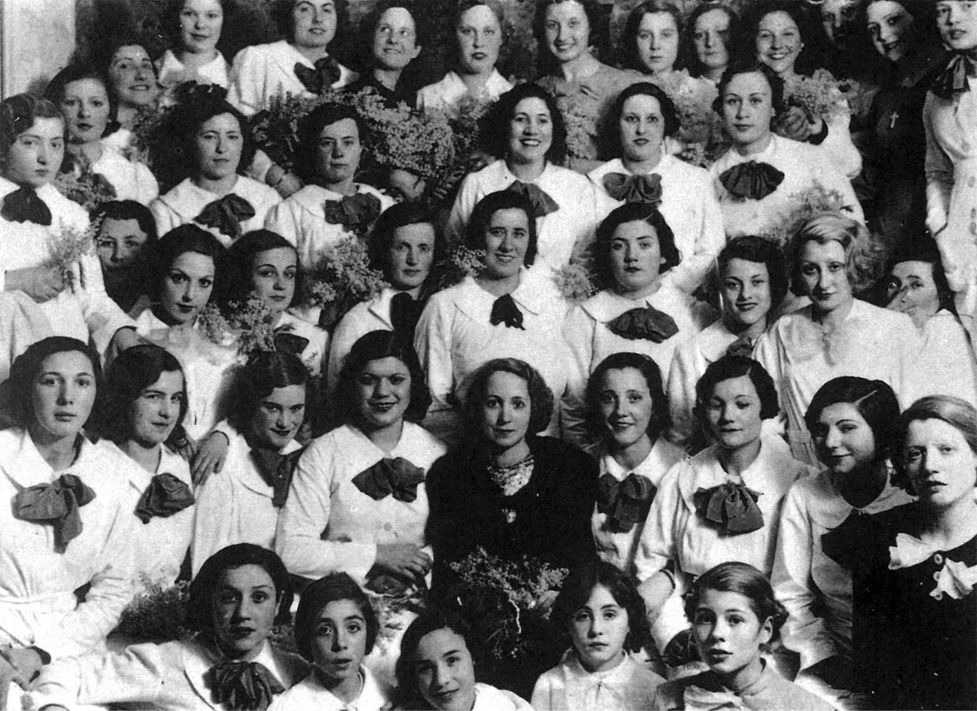 Black-and-white group portrait of women wearing matching white uniforms with dark bows. The image captures a formal gathering, possibly a school or organizational group, from an early 20th-century setting. The women hold flowers, adding a celebratory tone to the photograph.