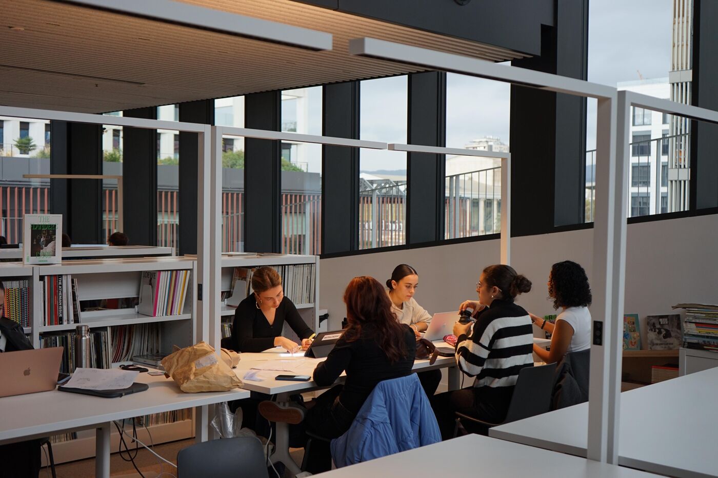 A group of students working together at a table in a modern library with large windows providing natural light. The shelves are filled with books and magazines, creating an academic and creative atmosphere. The scene emphasizes collaboration and learning.