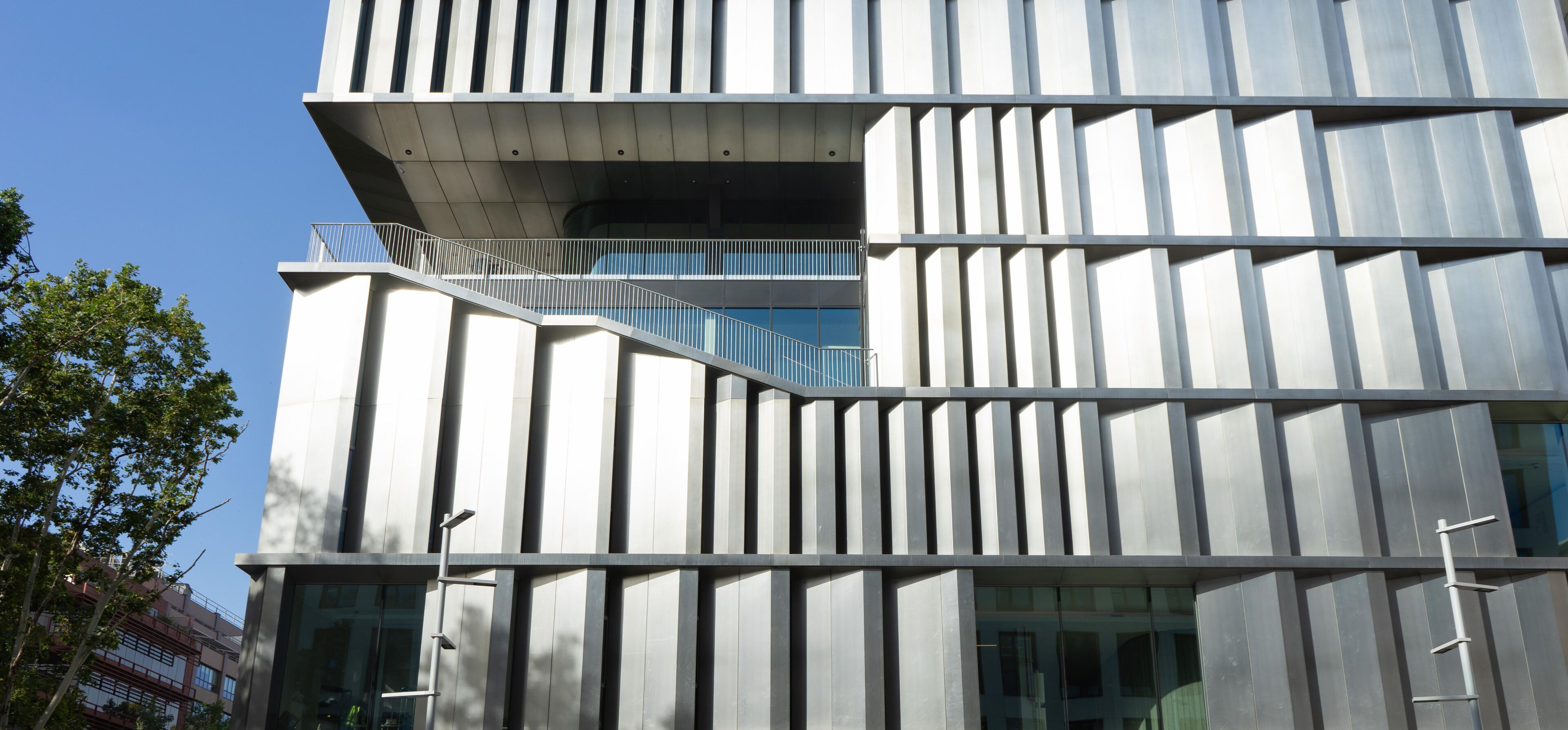 A close-up view of a modern building facade featuring vertical metallic panels and angular designs. The structure includes a stairway and balcony, with a tree adding a natural element. The contemporary style emphasizes geometry and clean lines.