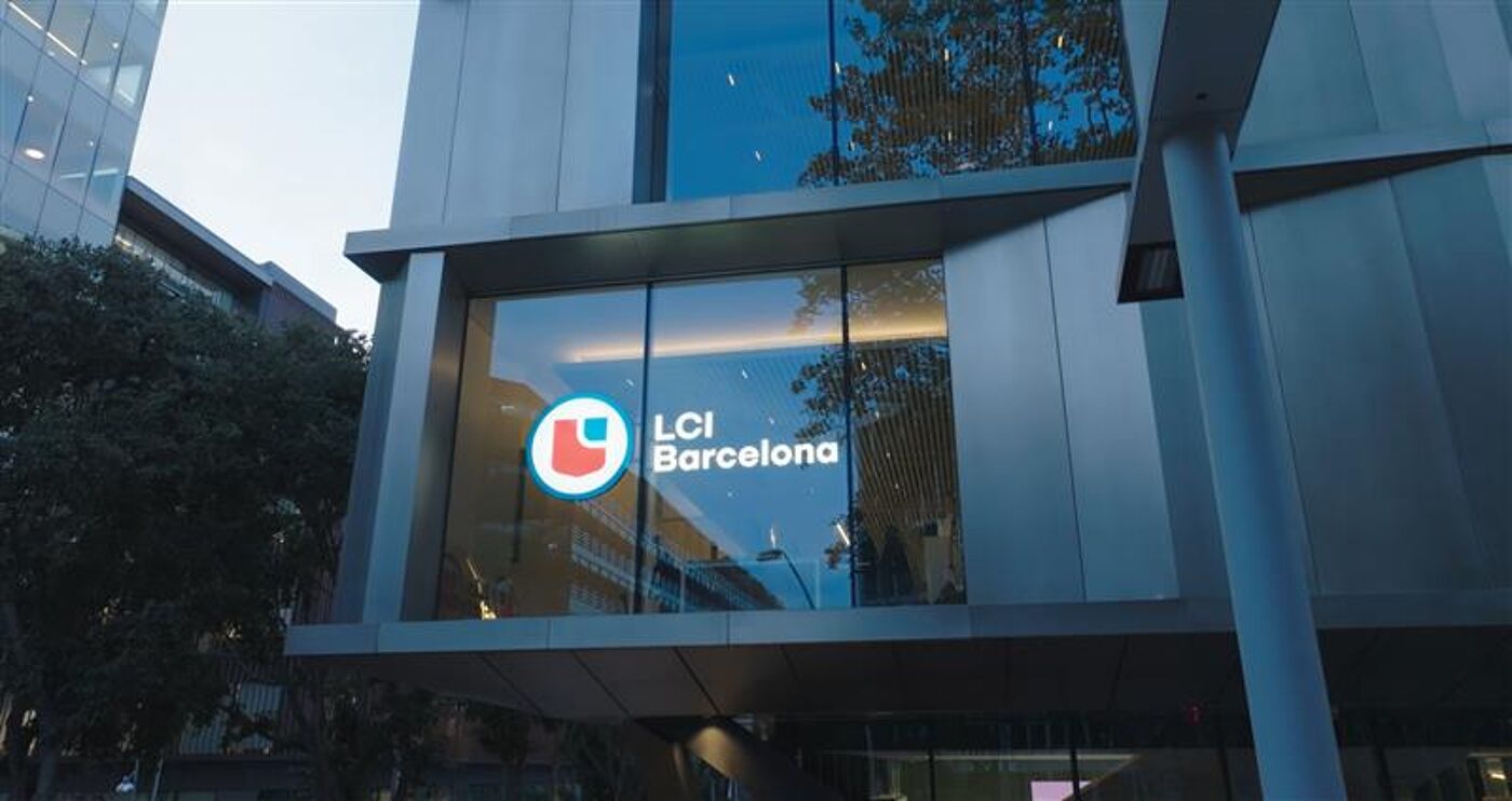 Glass-fronted facade of the LCI Barcelona building, featuring its illuminated logo and modern architectural design. The building is surrounded by urban greenery and reflects nearby structures, emphasizing a contemporary and professional aesthetic.