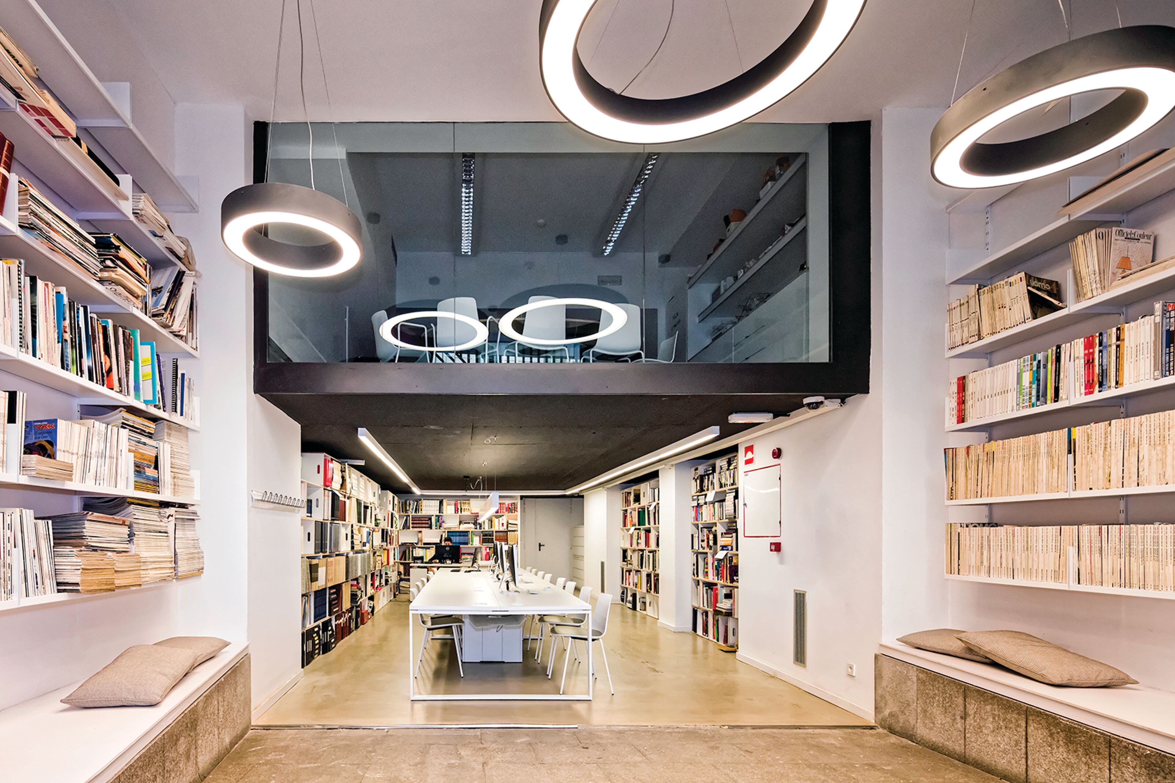 A sleek library space with book-lined walls and contemporary hanging lights, featuring a cozy reading area with a reflective ceiling for an open feel.