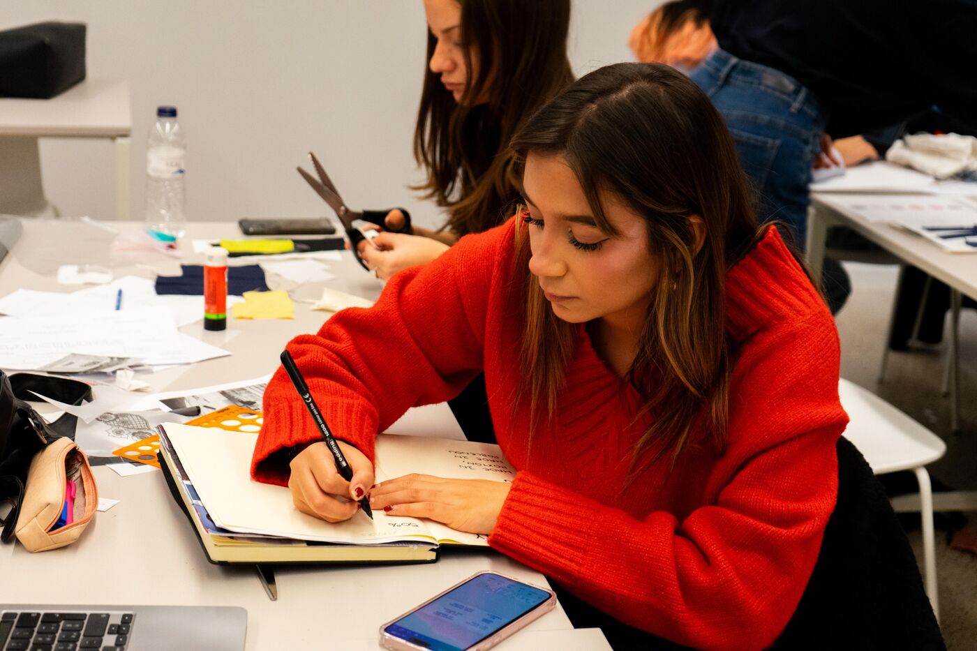 A young woman in a red sweater focuses on writing in a notebook while participating in a group creative activity. Scissors, paper, and other craft materials are scattered on the table, indicating an artistic or academic project.