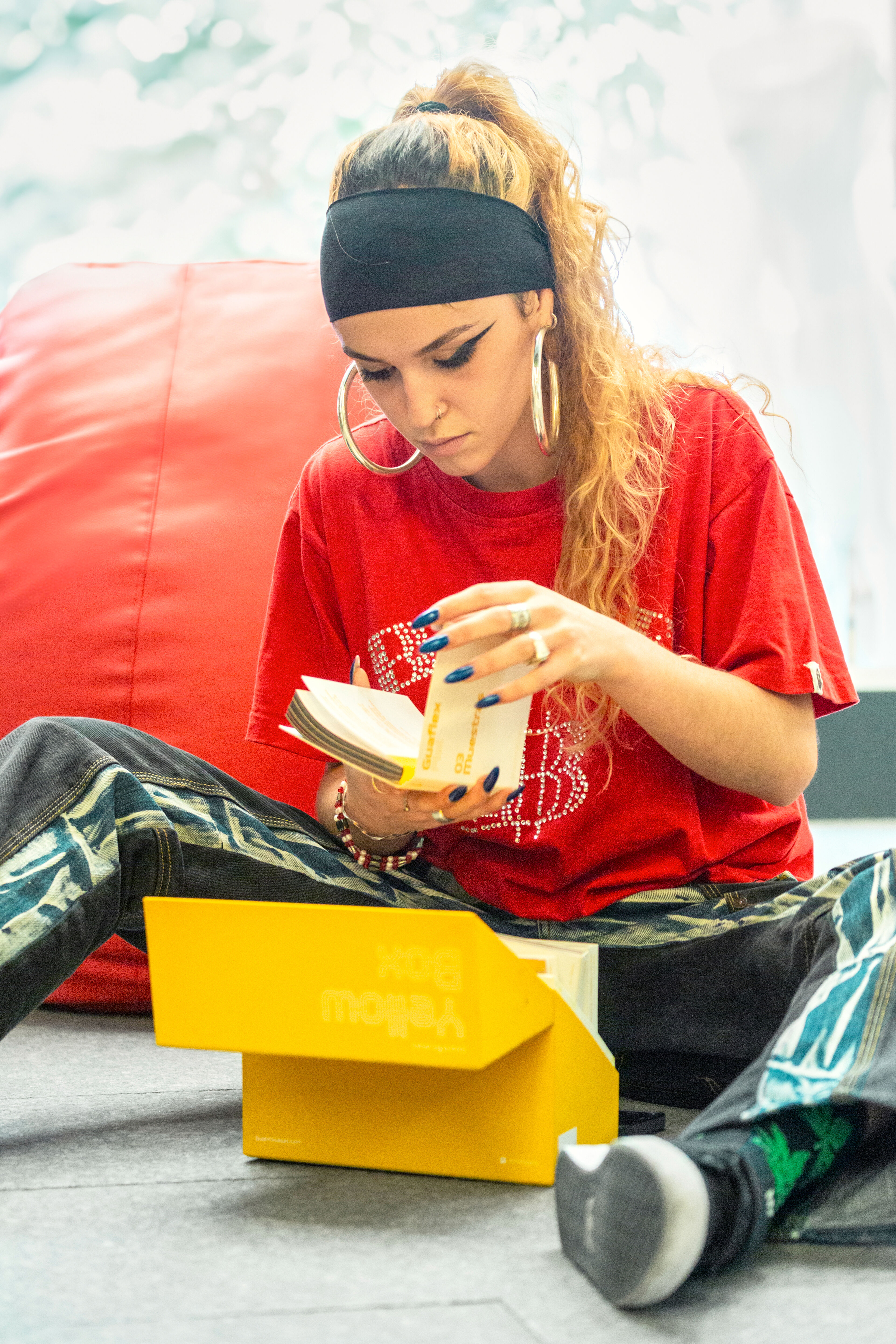  focused young woman in a red t-shirt and headband reads a book, sitting on the floor.