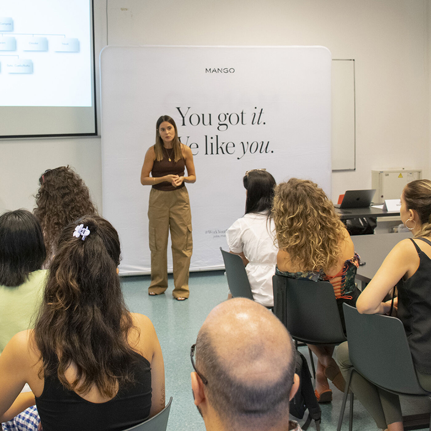 A speaker addresses an audience during a branding workshop, with a backdrop displaying the slogan "You got it. We like you." The presentation includes slides, and attendees appear attentive, creating a focused and professional atmosphere.