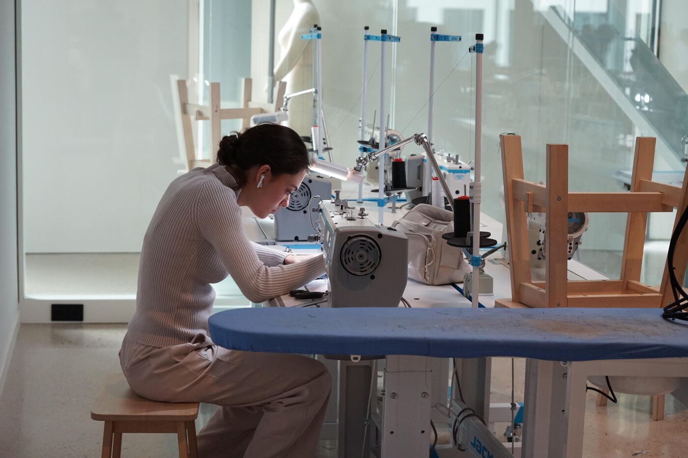 A woman focuses intently on operating a sewing machine in a bright and modern workshop. She wears earbuds and sits at a table surrounded by other sewing machines and tools, suggesting a professional or educational setting for garment creation.