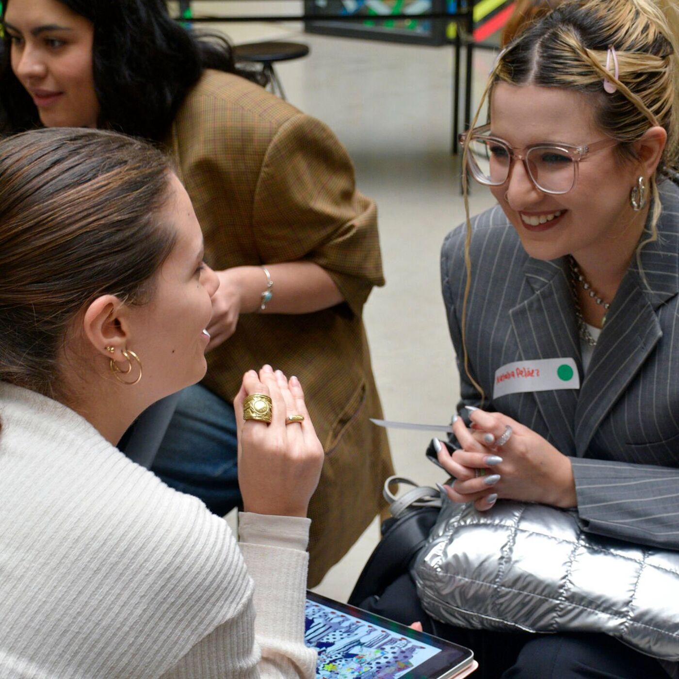 Three women engaged in a friendly conversation, with one holding a tablet displaying colorful imagery. They appear to be part of a collaborative or networking event, with name tags indicating introductions. The atmosphere is informal yet professional.