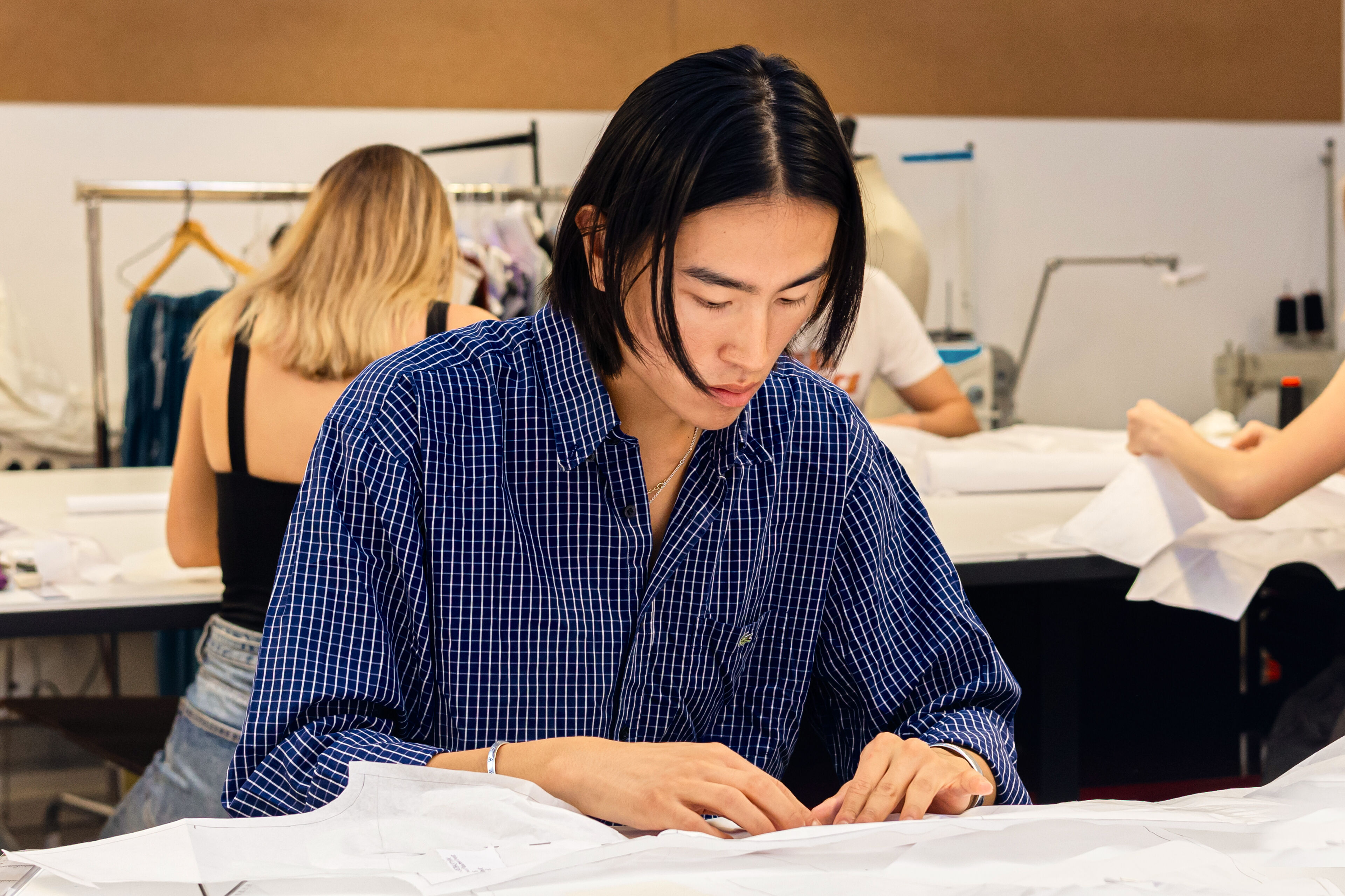 A diligent fashion student in a blue checked shirt, engrossed in drafting a pattern.
