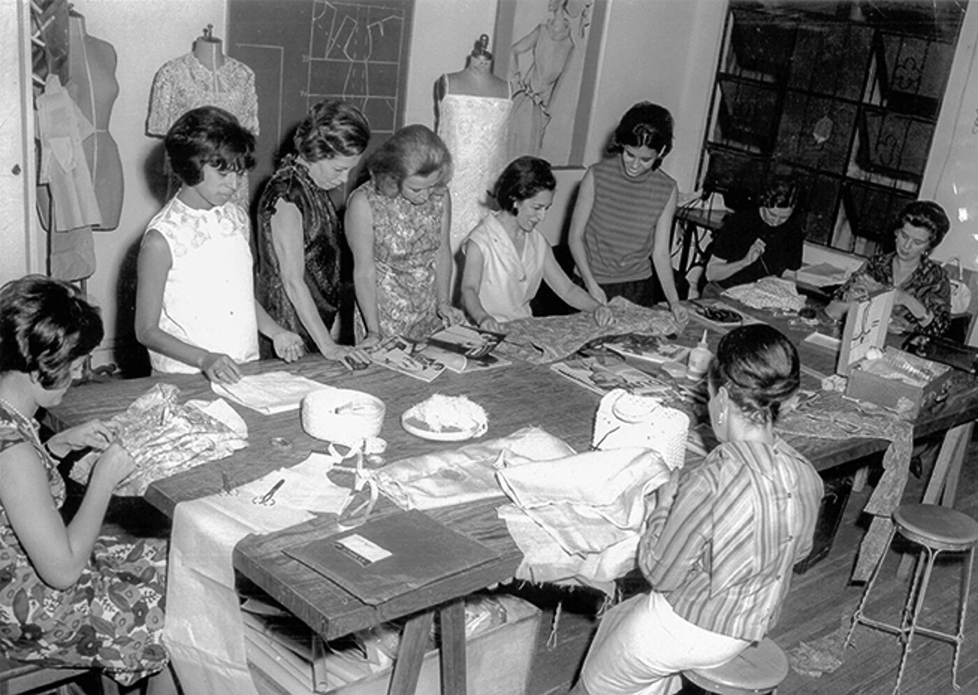 black and white photograph depicting eight women engaged in sewing activities around a large table, with mannequins and fabric in the background.