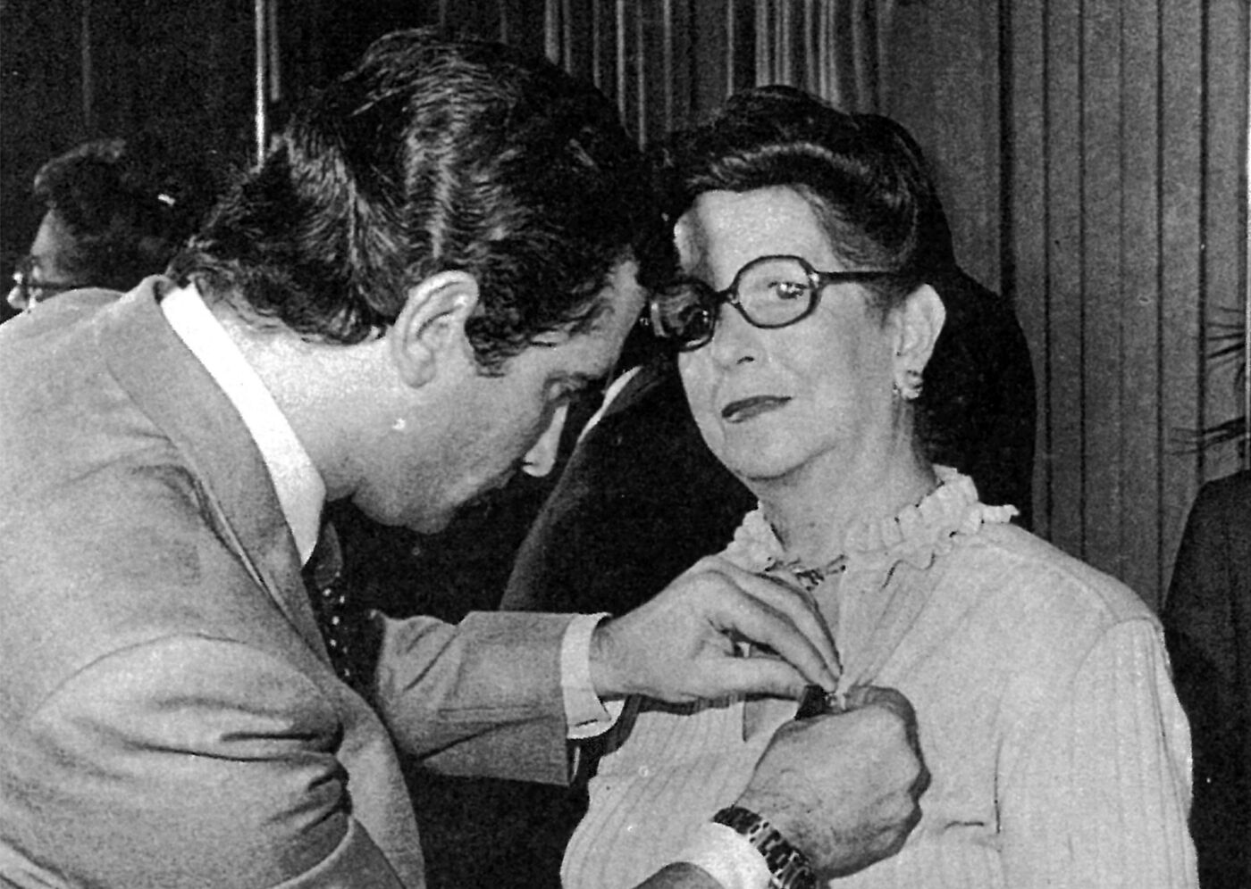 In the picture, we have a man and a woman standing next to each other. The man is fixing a pin or a brooch on the woman's clothing, possibly her neck or her dress. The woman is wearing glasses, and the man is adjusting the pin to ensure it is properly placed and secure.