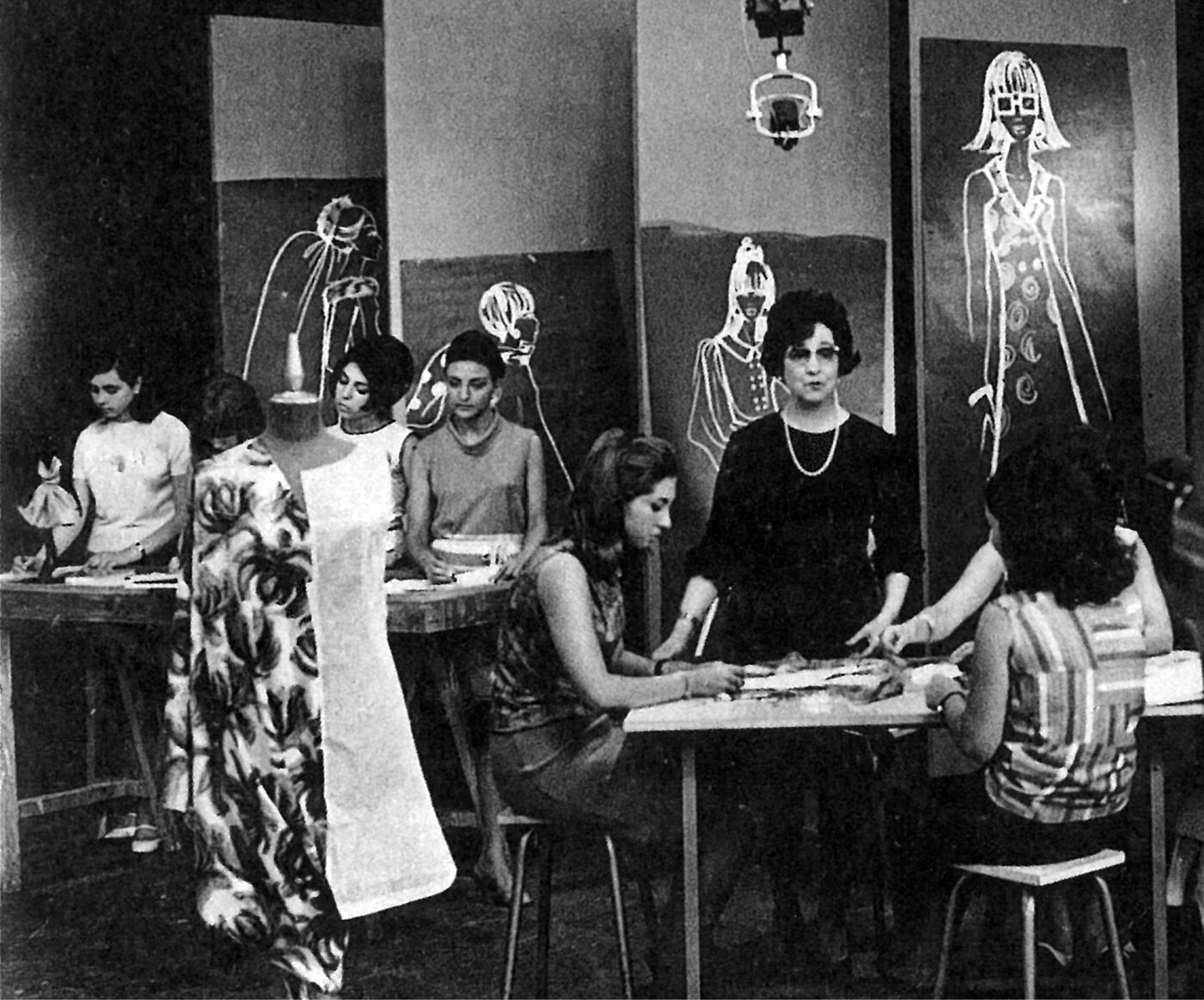 The image shows a group of women in what appears to be a fashion design class, with sketches of outfits on display and mannequins dressed in fabric.