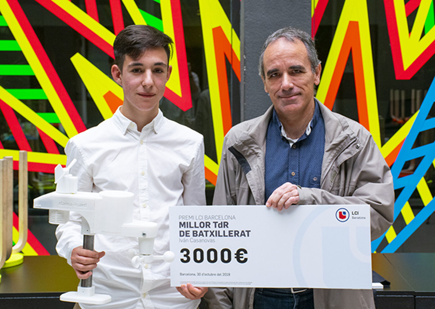  A young individual and an older man pose with a celebratory check for €3000, awarded for the 'Best Baccalaureate Thesis' in Barcelona, dated October 2019.